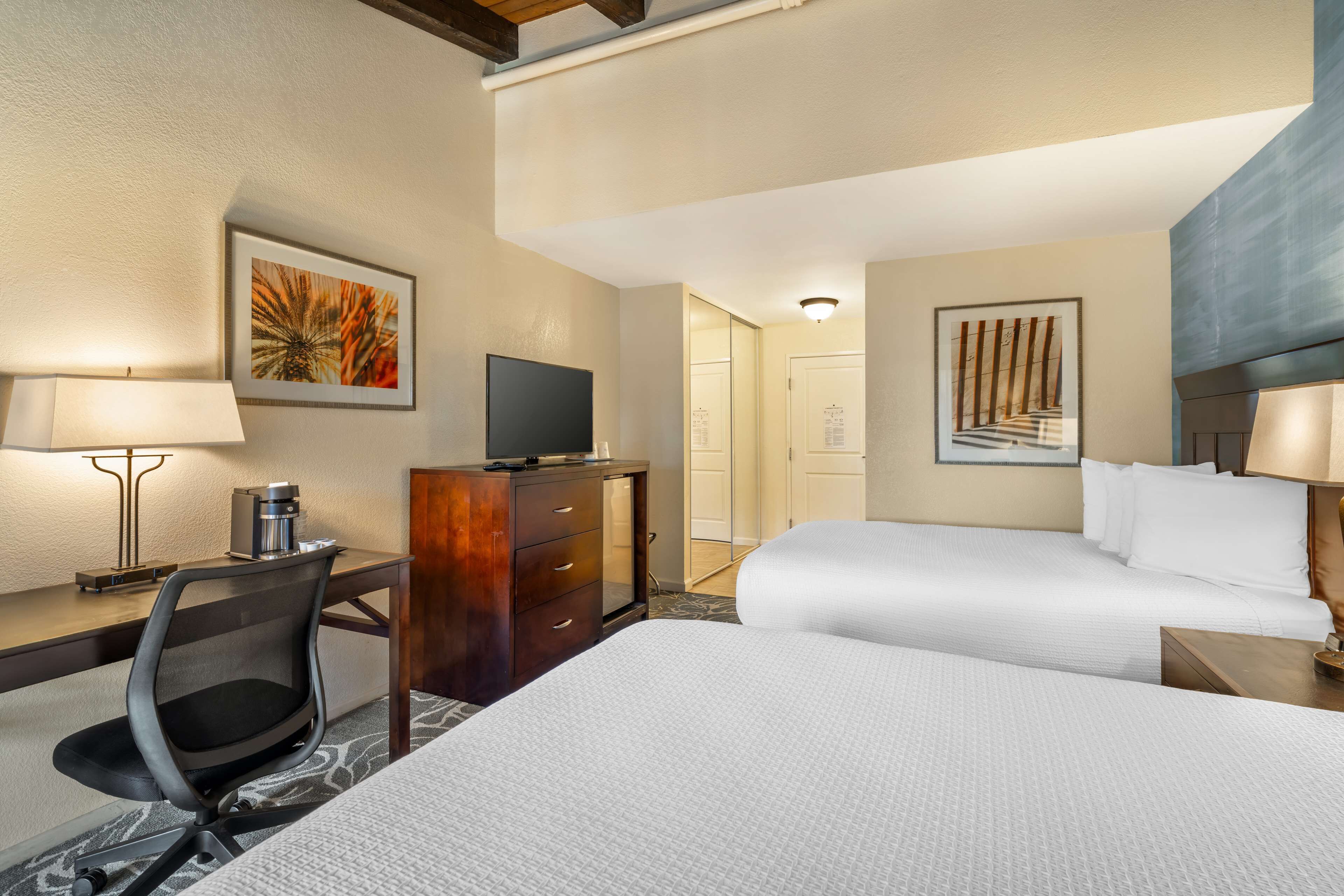 Habitación de hotel con dos camas, un escritorio con silla, televisor y obras de arte en las paredes.