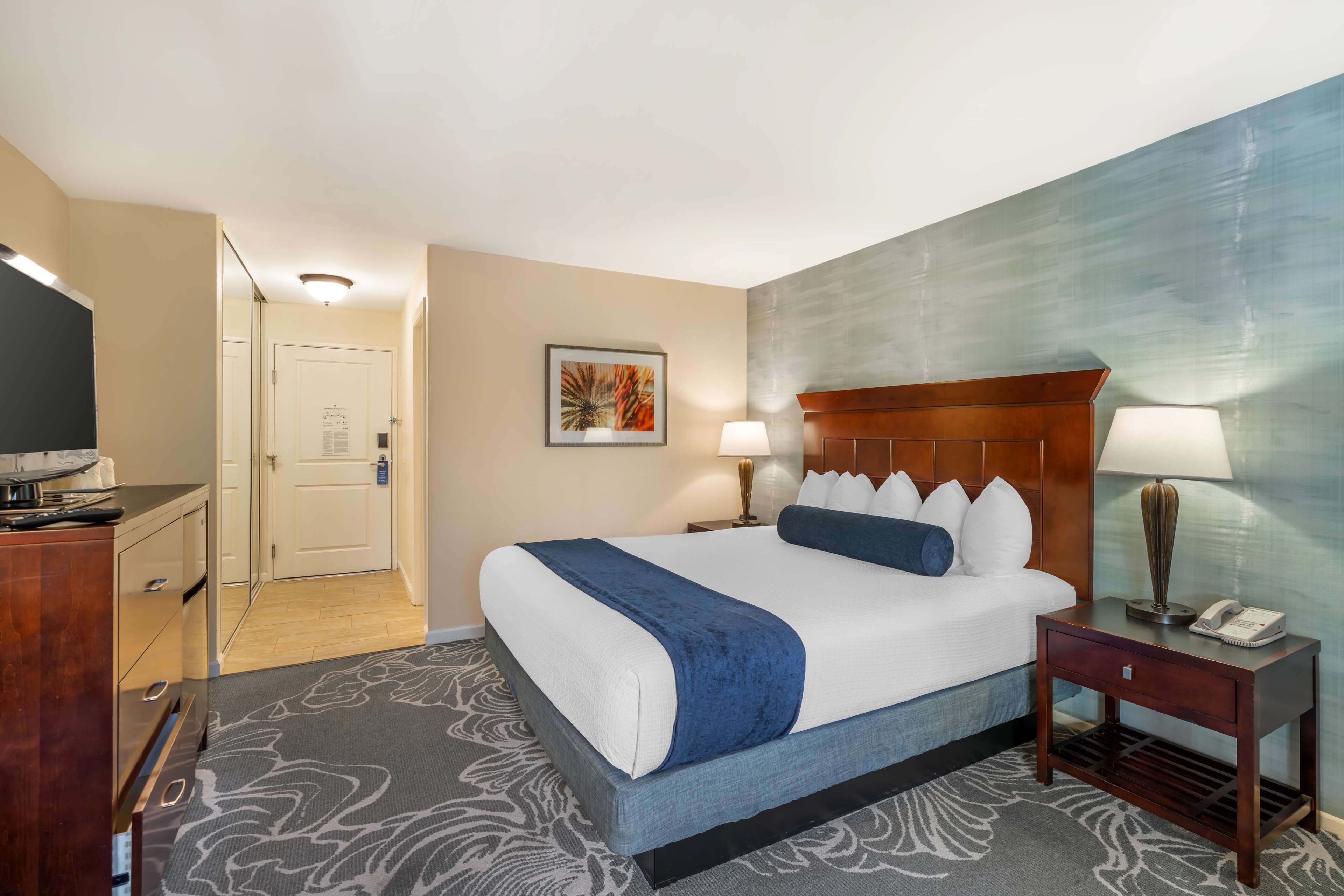 A hotel room with a large bed, two bedside tables with lamps, a TV, and a door leading to a hallway.