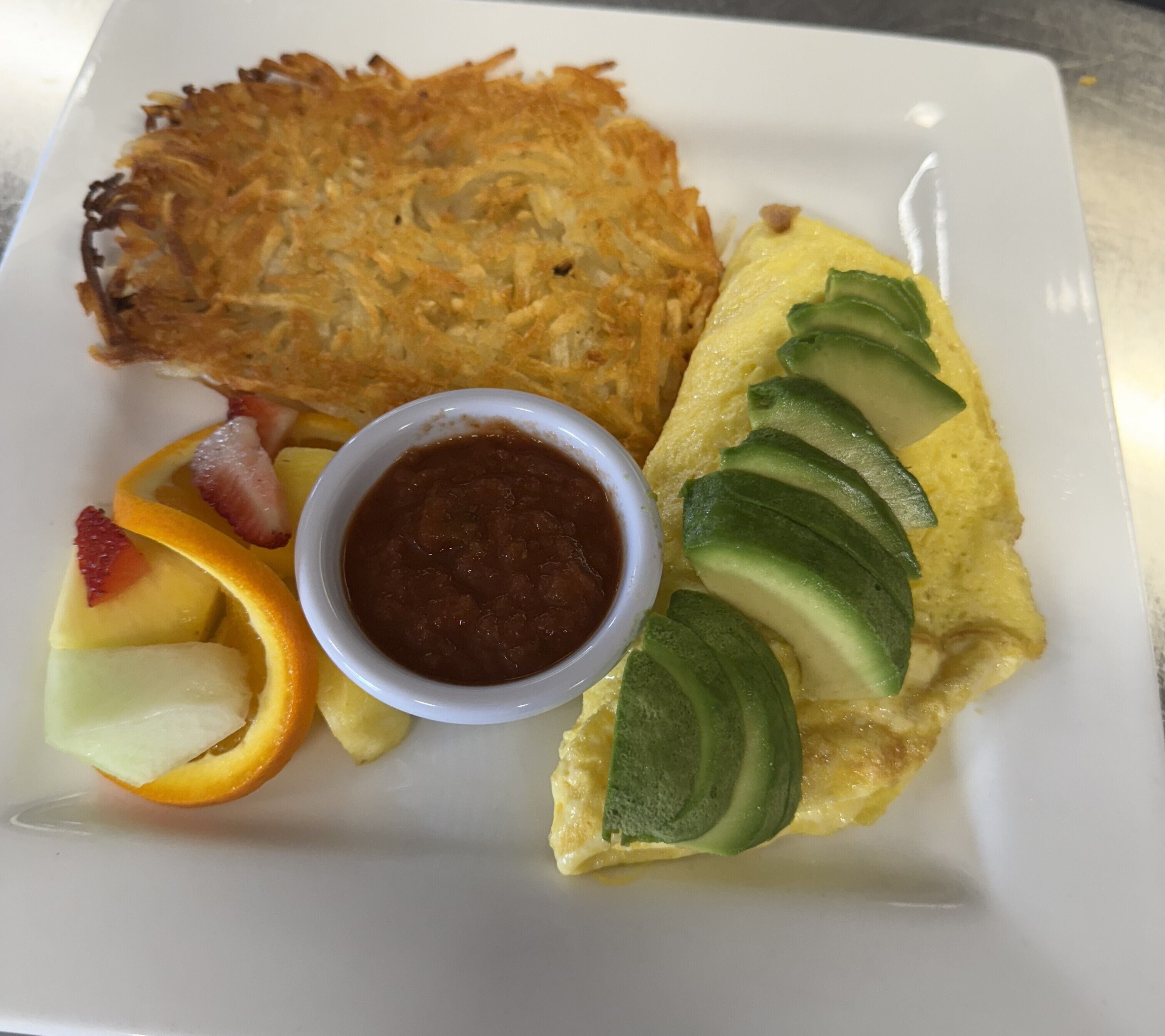 A plate with a hash brown, omelette, sliced fruits, and a small bowl of sauce.