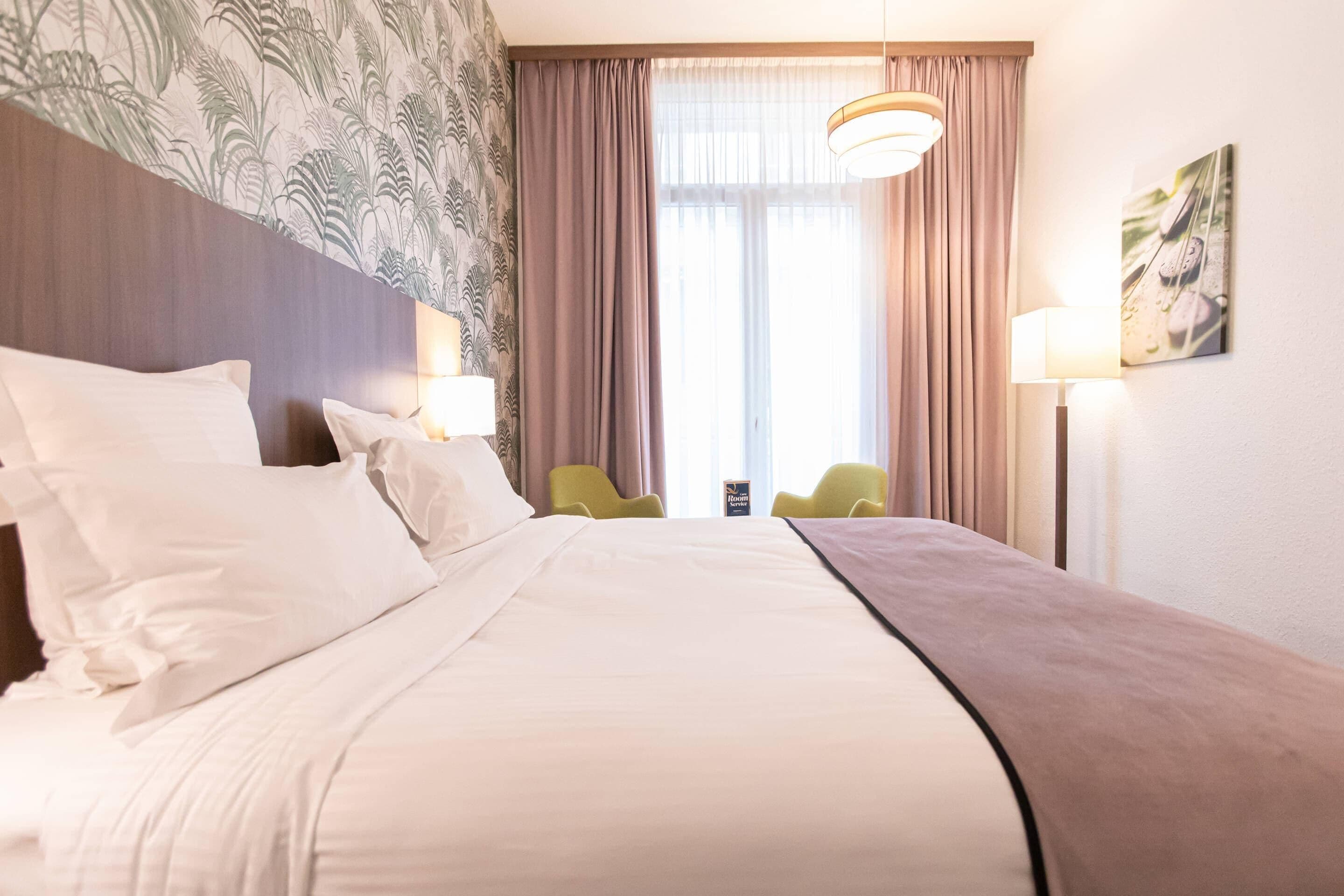 Habitación de hotel con una cama grande, dos lámparas de noche, una ventana con cortinas, una silla verde y un cuadro en la pared.