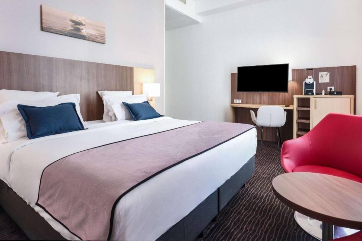 A hotel room with a bed, a desk, a TV, a red chair, and a small table.