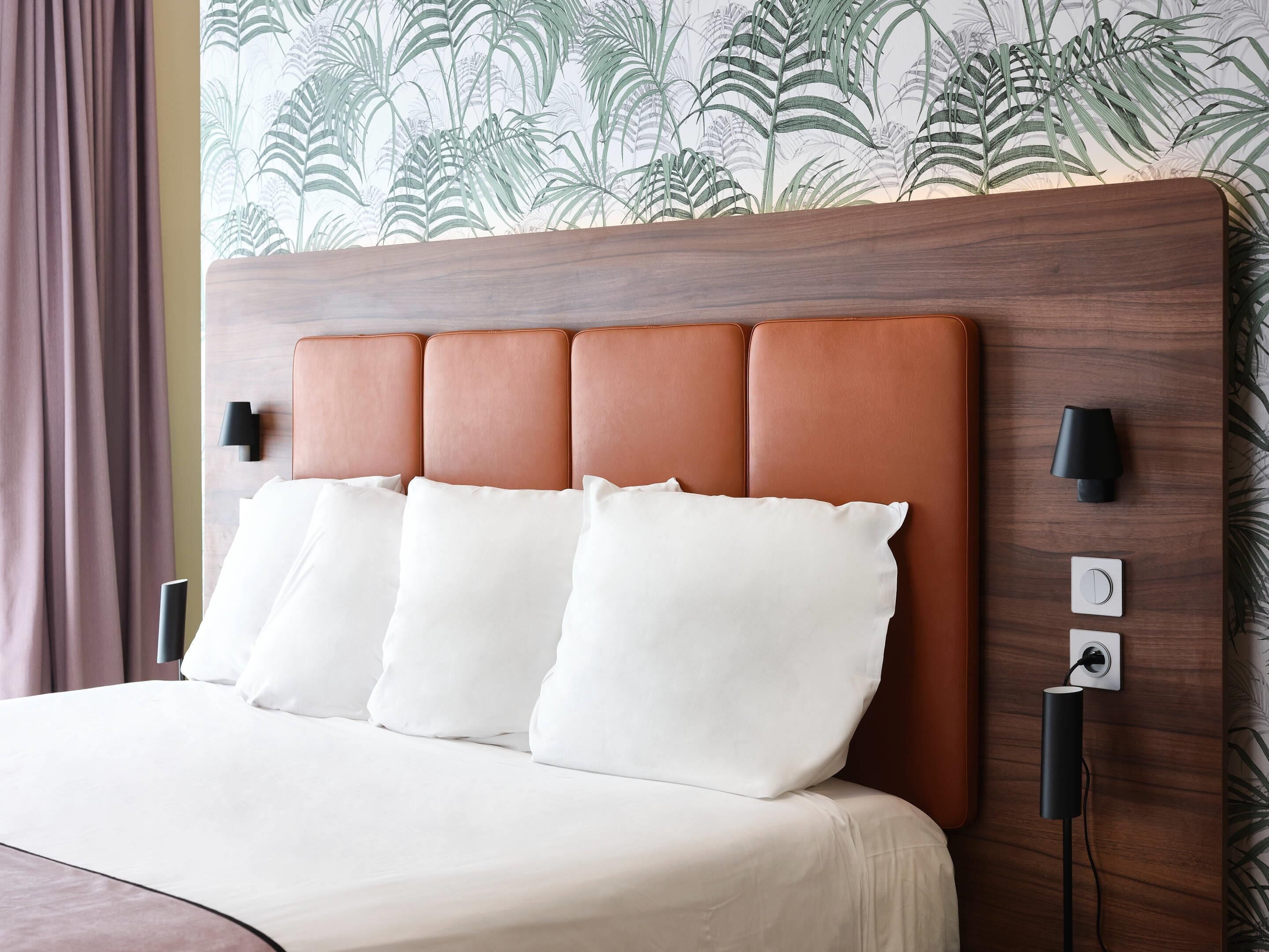 A hotel room with a bed, a headboard, a wall-mounted lamp, and a power outlet.
