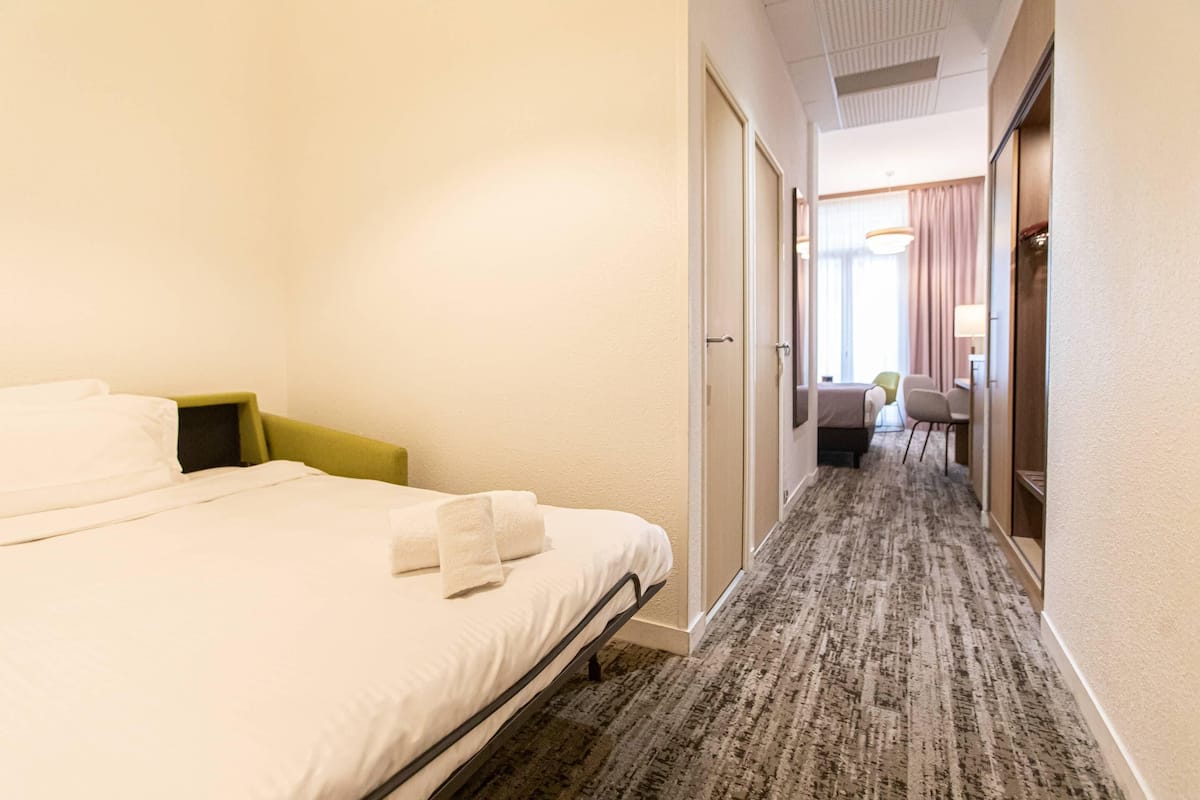 A hotel room with a bed, a bedside table, a chair, and a desk with a lamp.