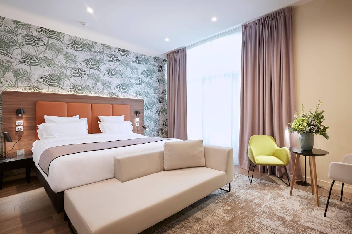 A hotel room with a large bed, a sofa, a chair, a small table, and a potted plant.