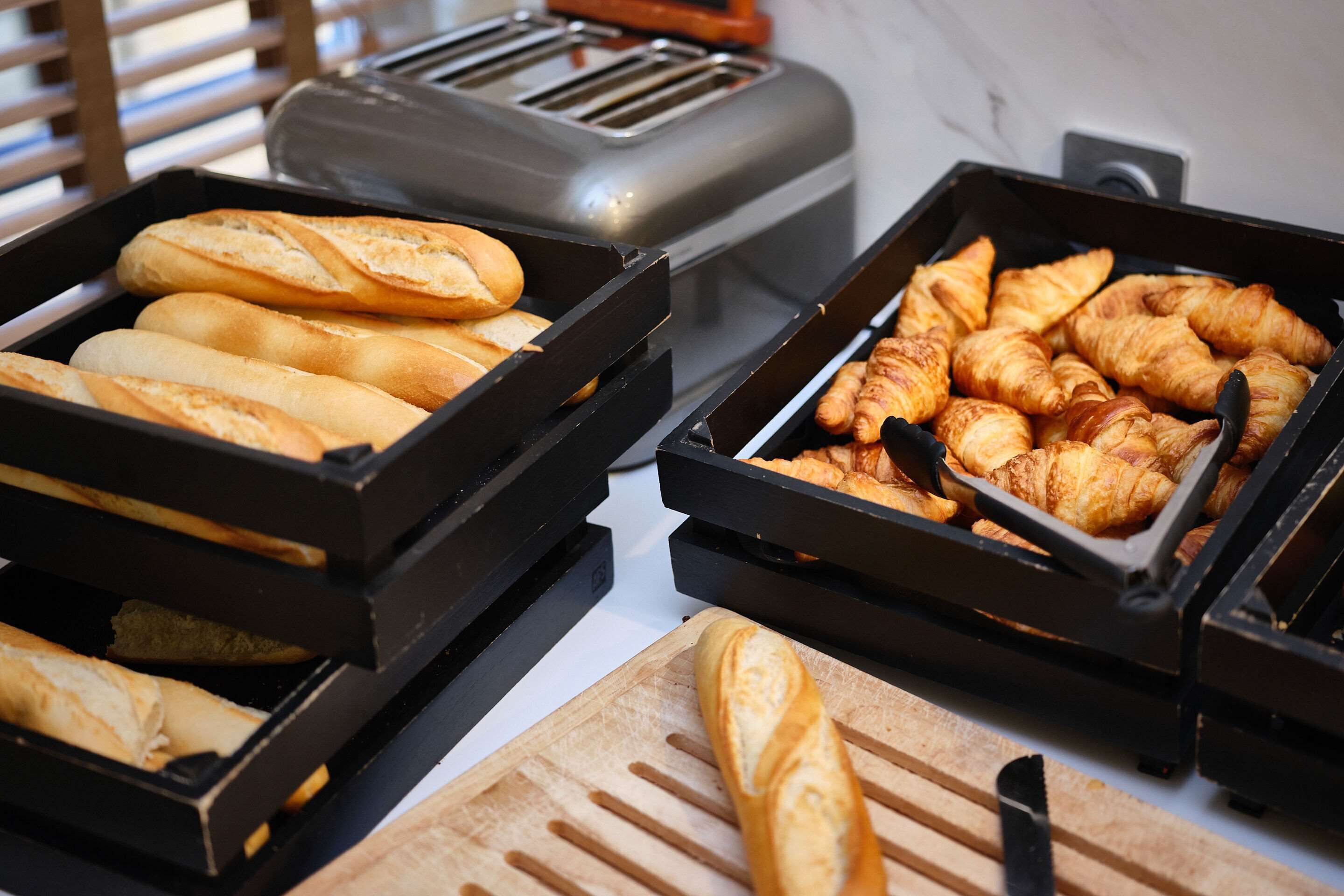 Two black trays of bread, one with baguettes and the other with croissants, on a countertop.