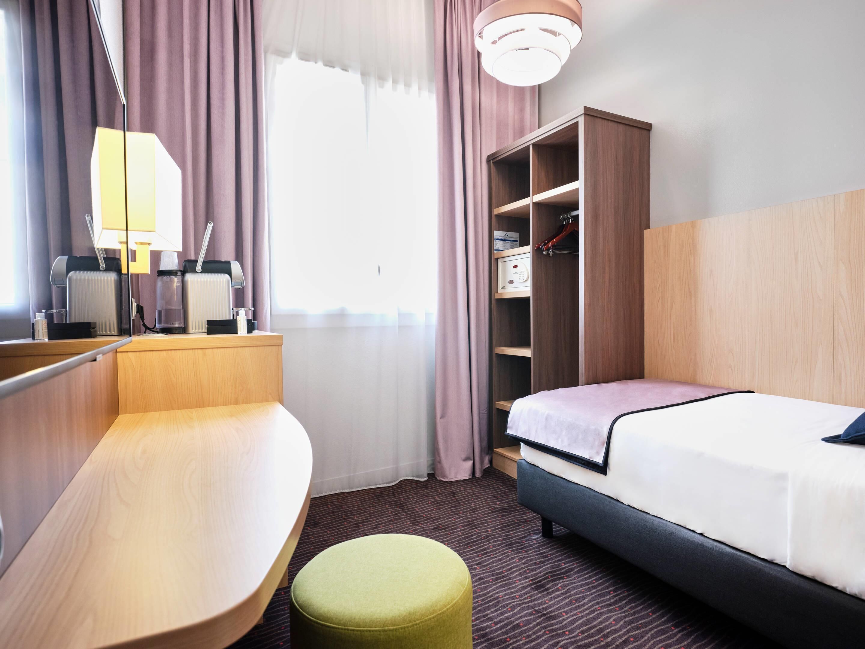A hotel room with a bed, a desk, a chair, a small stool, a wardrobe, and a window with curtains.
