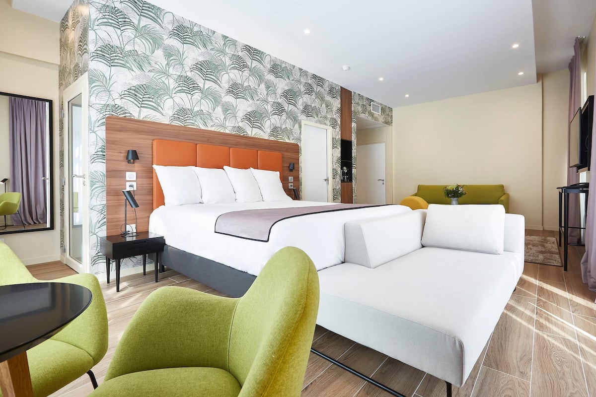 A hotel room with a bed, green armchairs, a white sofa, a small table, and a wall with a leaf pattern.