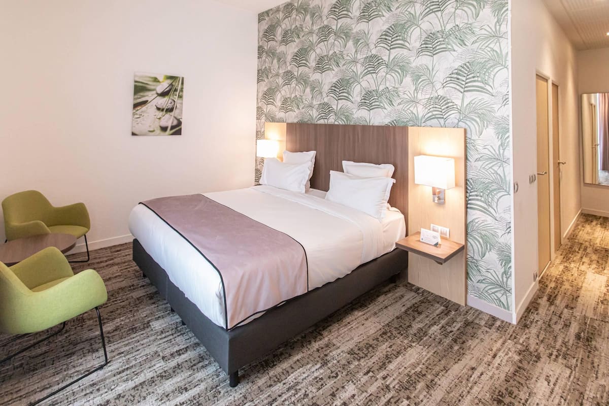 A hotel room with a bed, two bedside lamps, a green chair, and a patterned wallpaper.