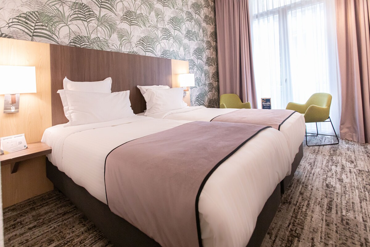 Une chambre d’hôtel avec deux lits, une tête de lit, une table de chevet, une lampe, une chaise et une fenêtre avec des rideaux.