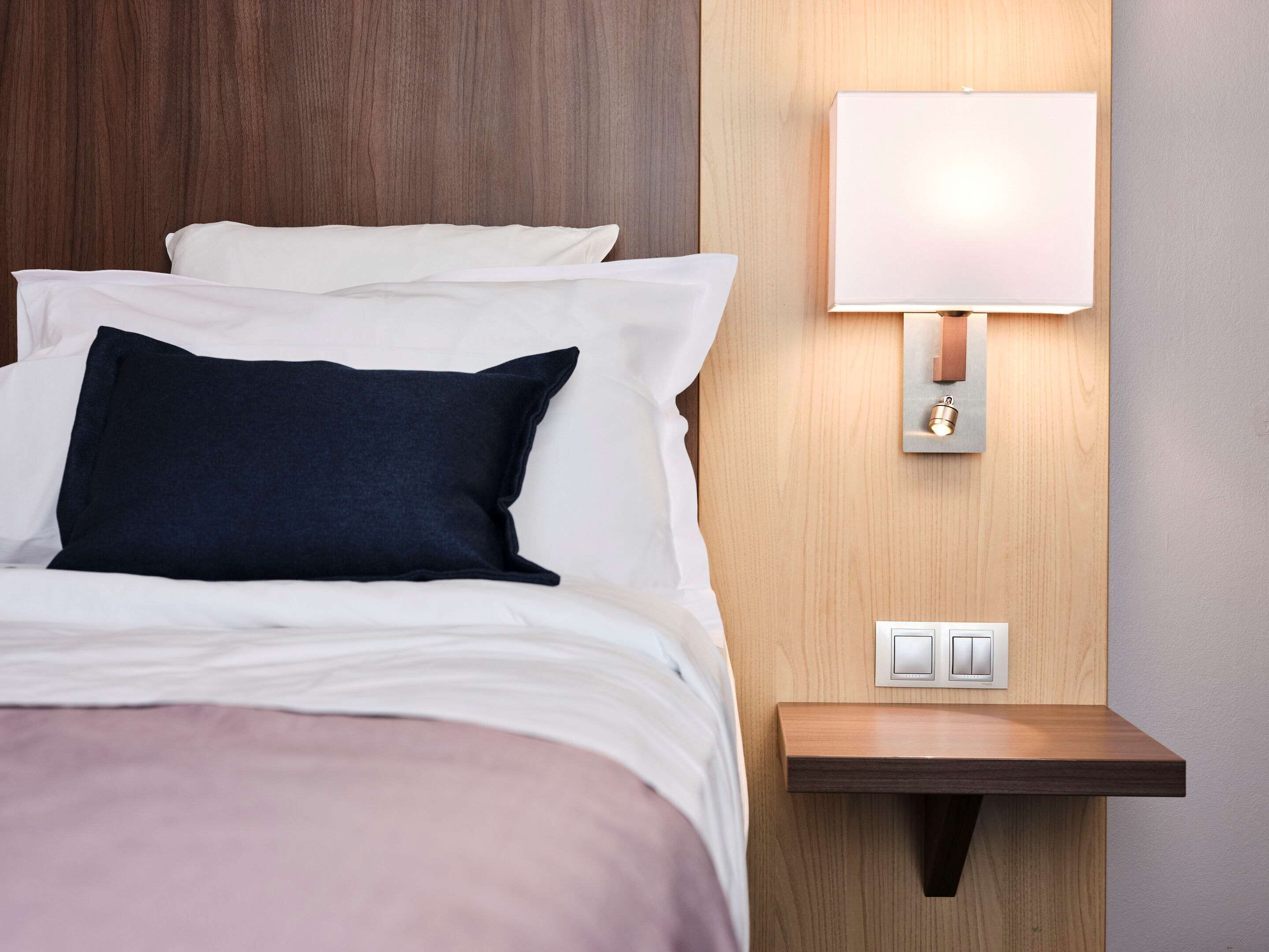 A neatly made bed with white and dark blue pillows, a bedside table with a lamp, and a wall-mounted light switch.