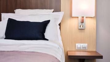 A neatly made bed with white and dark blue pillows, a bedside table with a lamp, and a wall-mounted light switch.