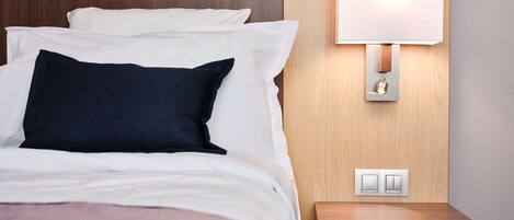 Una cama bien tendida con almohadillas blancas y azul oscuro, una mesita de noche con lámpara, e interruptor de luz en la pared.