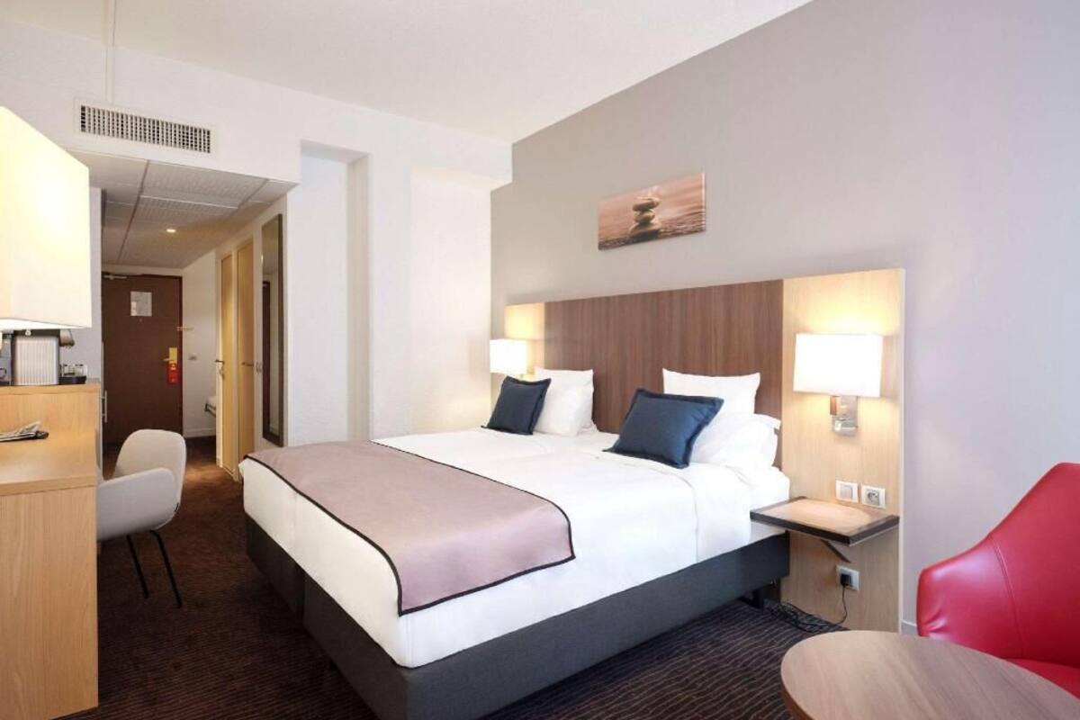 A hotel room with a bed, bedside tables, a desk, a chair, and a red chair.