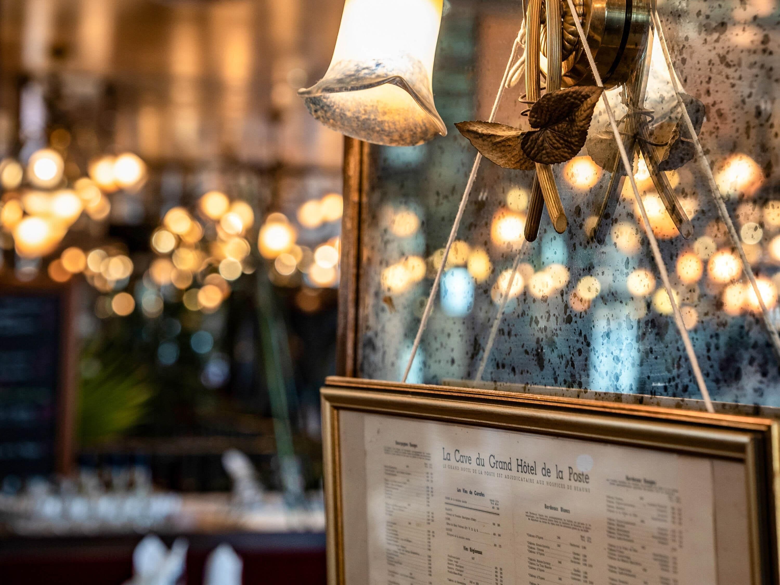 A menu board for La Cave du Grand Hotel de la Poste with a blurred background of lights.