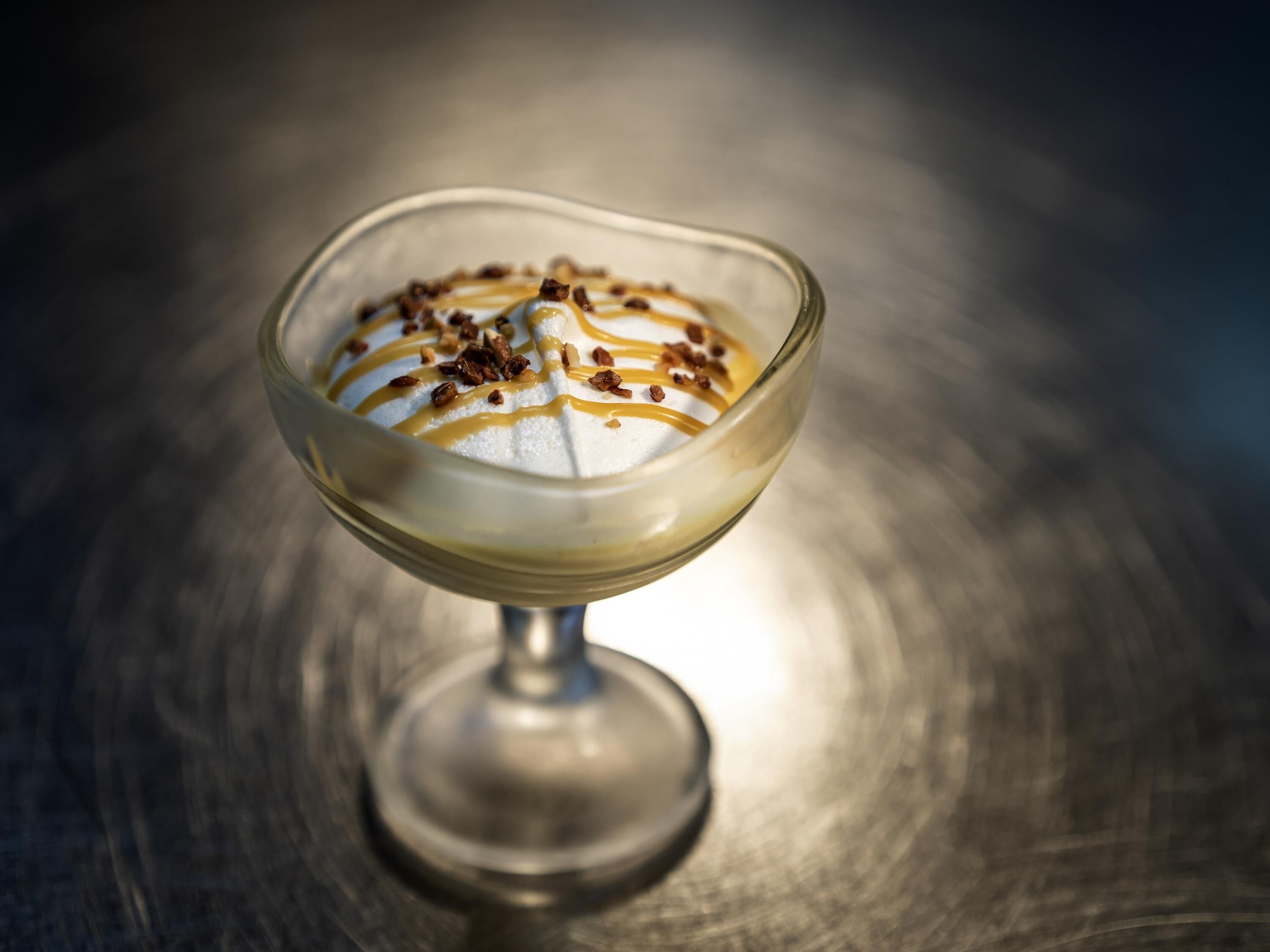 Een dessert in een glazen schaal met karamelsaus en noten op een reflecterend oppervlak.