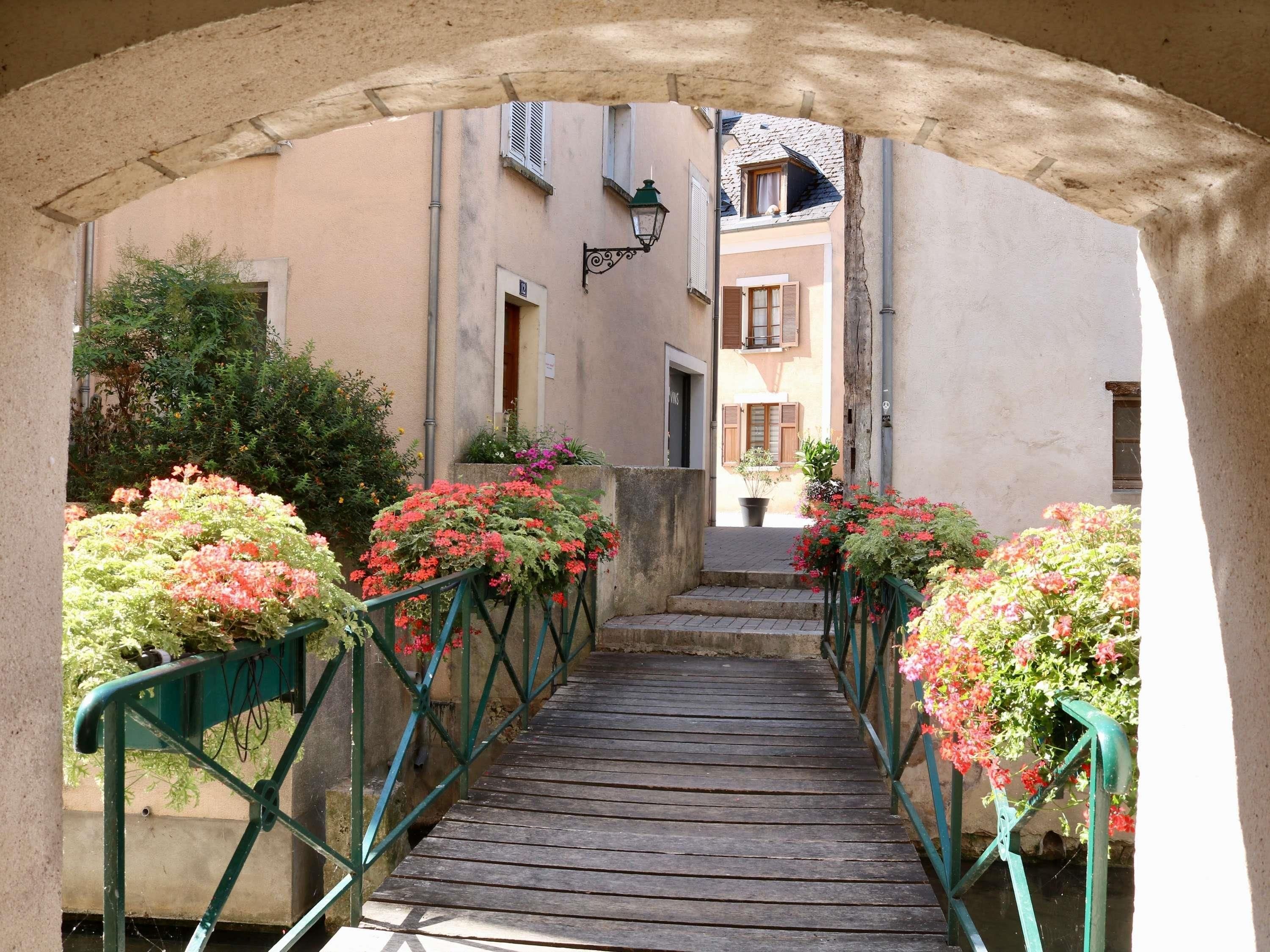 Une ruelle étroite avec un pont en bois, bordée de verdure et de bâtiments aux volets fermés.