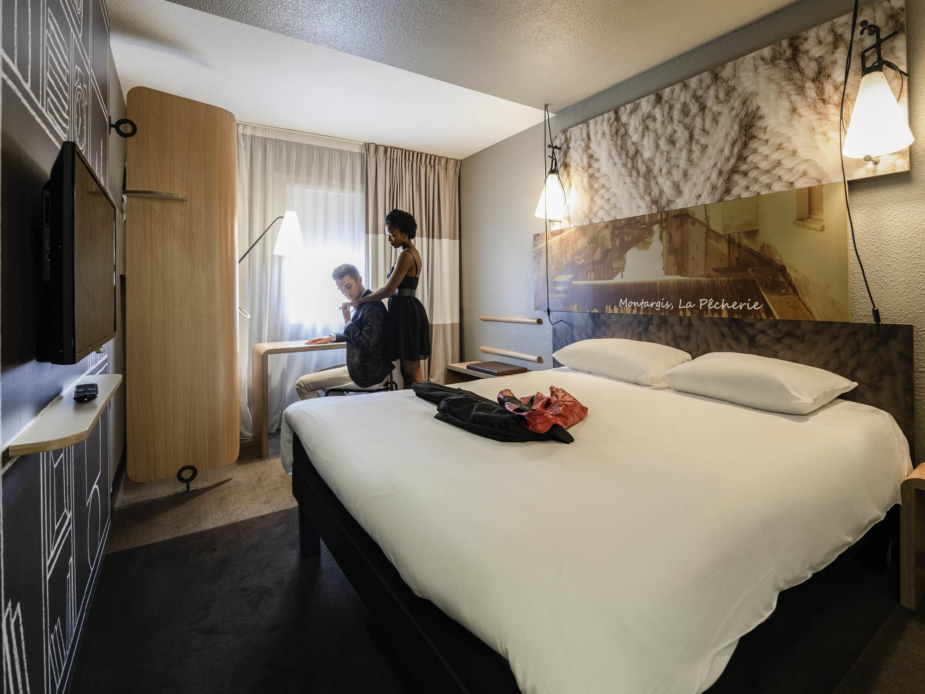 A hotel room with a bed, a desk, a chair, a TV, and a large mirror.