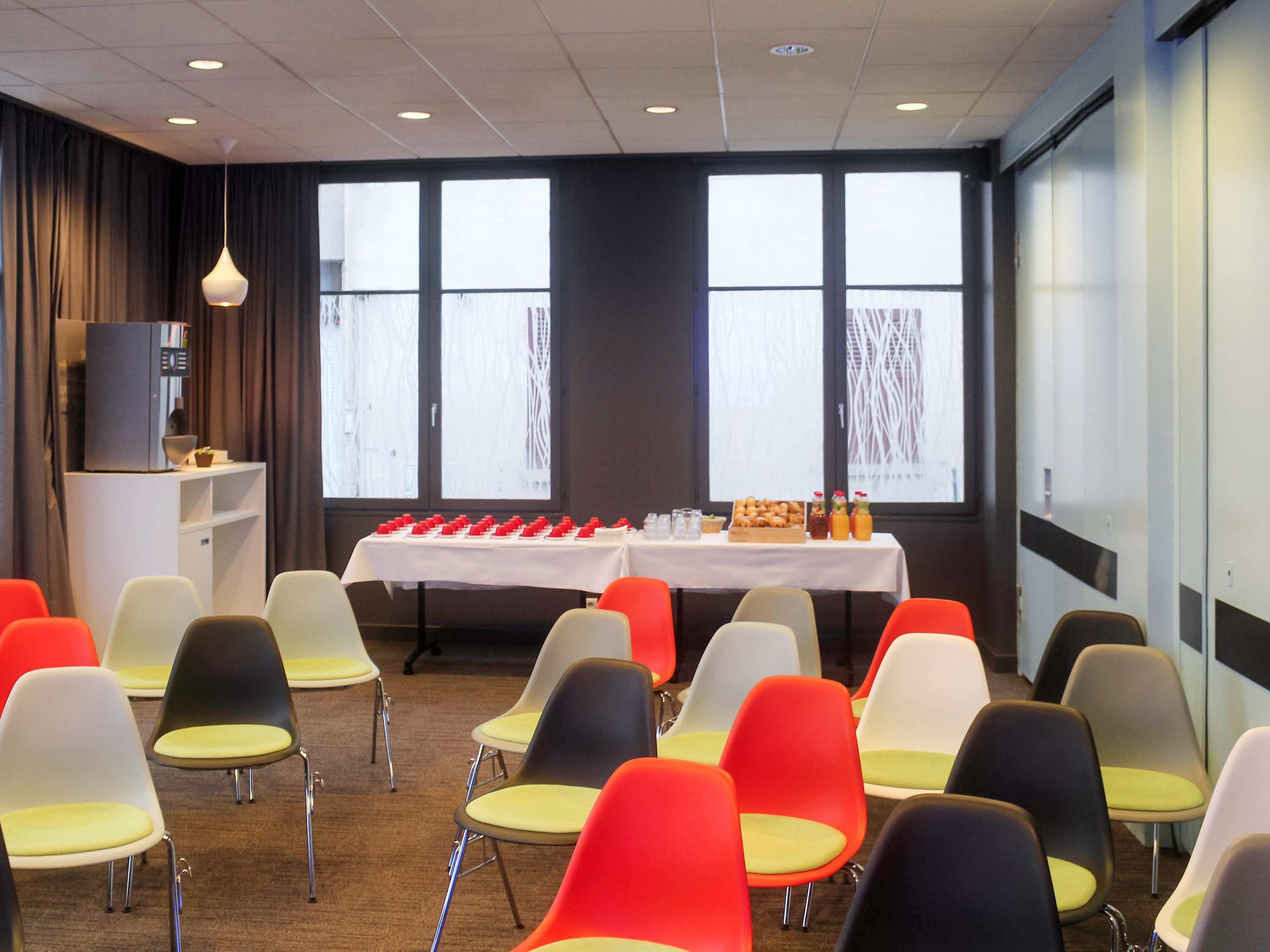 Un salón de conferencias con filas de sillas, una mesa con refrigerios y ventanas amplias.