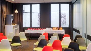 Un salón de conferencias con filas de sillas, una mesa con refrigerios y ventanas amplias.