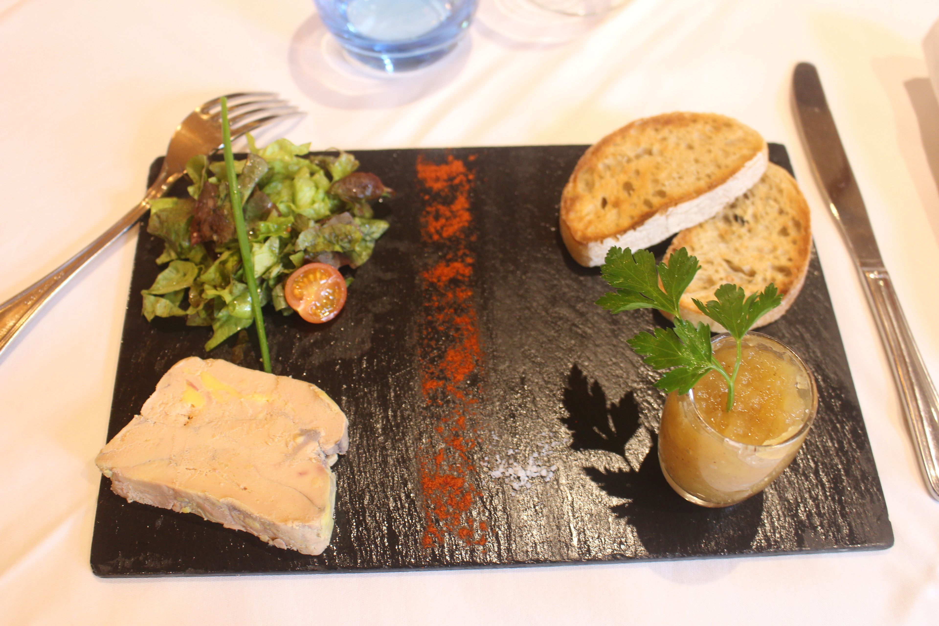 A black slate board with a piece of pâté, a salad, two slices of bread, and a small glass of sauce.