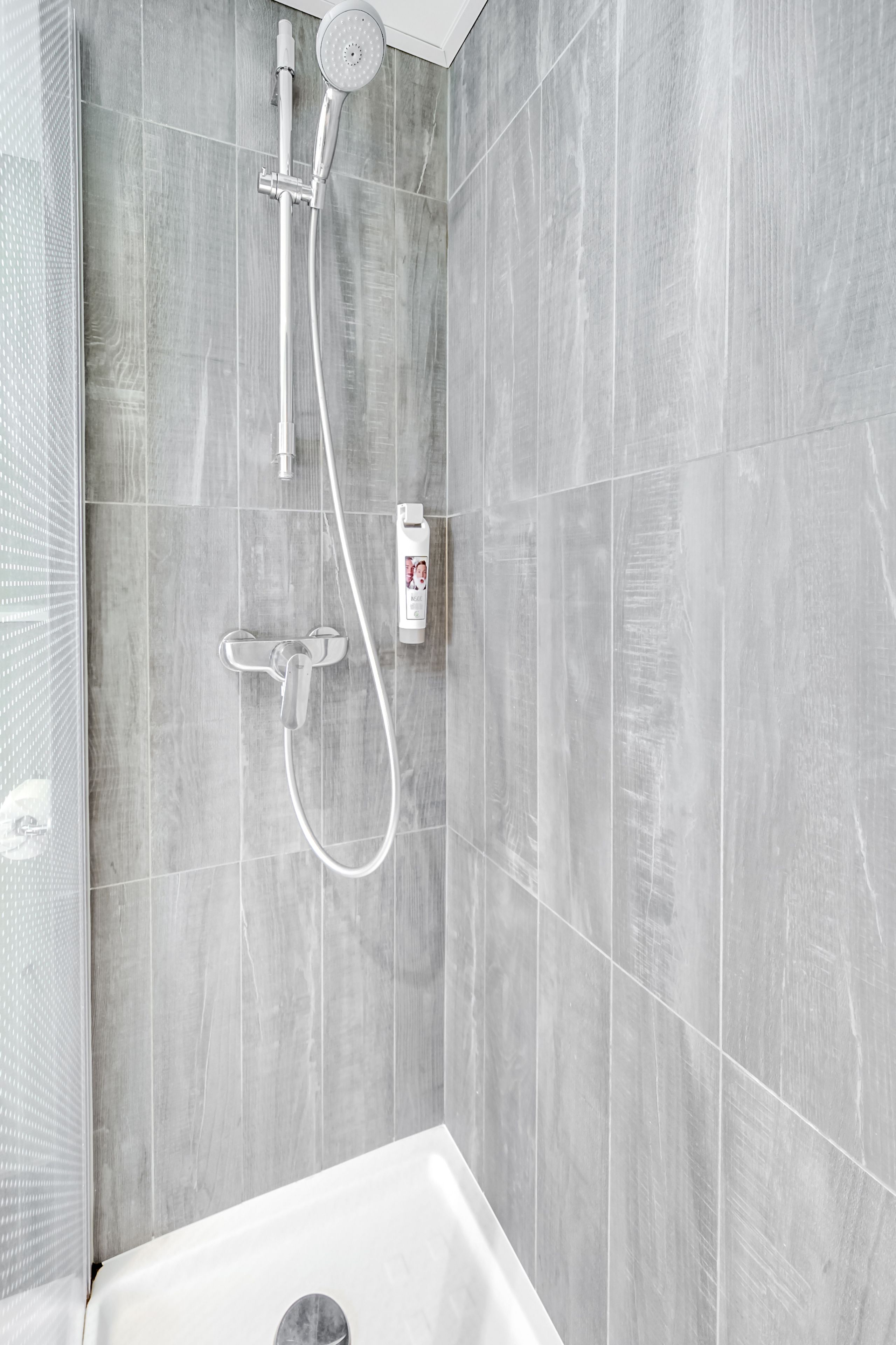 A modern bathroom with grey marble tiles, a shower with a handheld showerhead and a handheld shower attachment, and a bottle of shampoo on the wall.