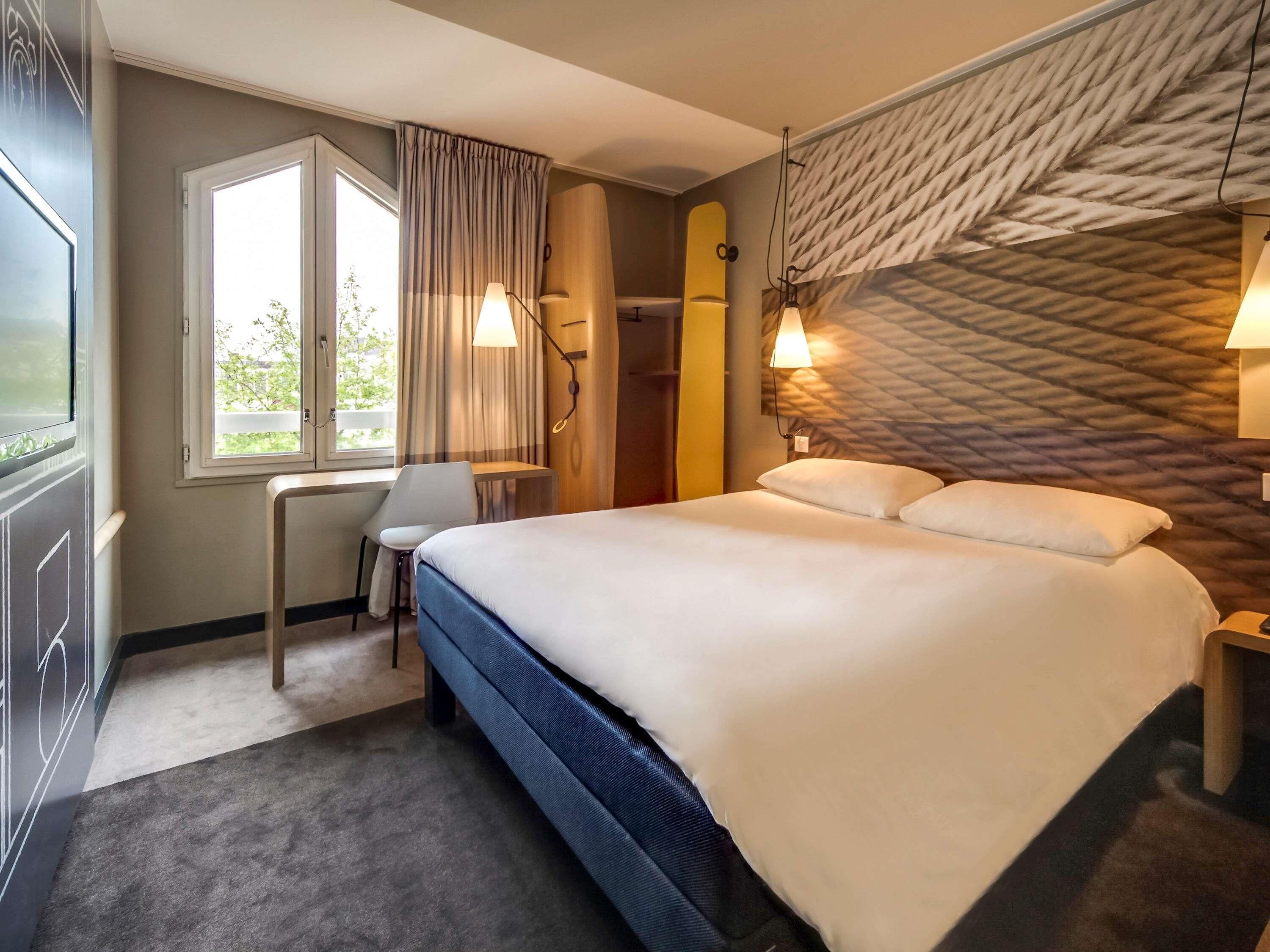A hotel room with a bed, a desk, a chair, a window, and a wall-mounted lamp.