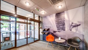 A modern interior with a glass door entrance, a seating area with two chairs, a small table, and a large wall mural. - ibis Orléans Centre Gare (Orléans)