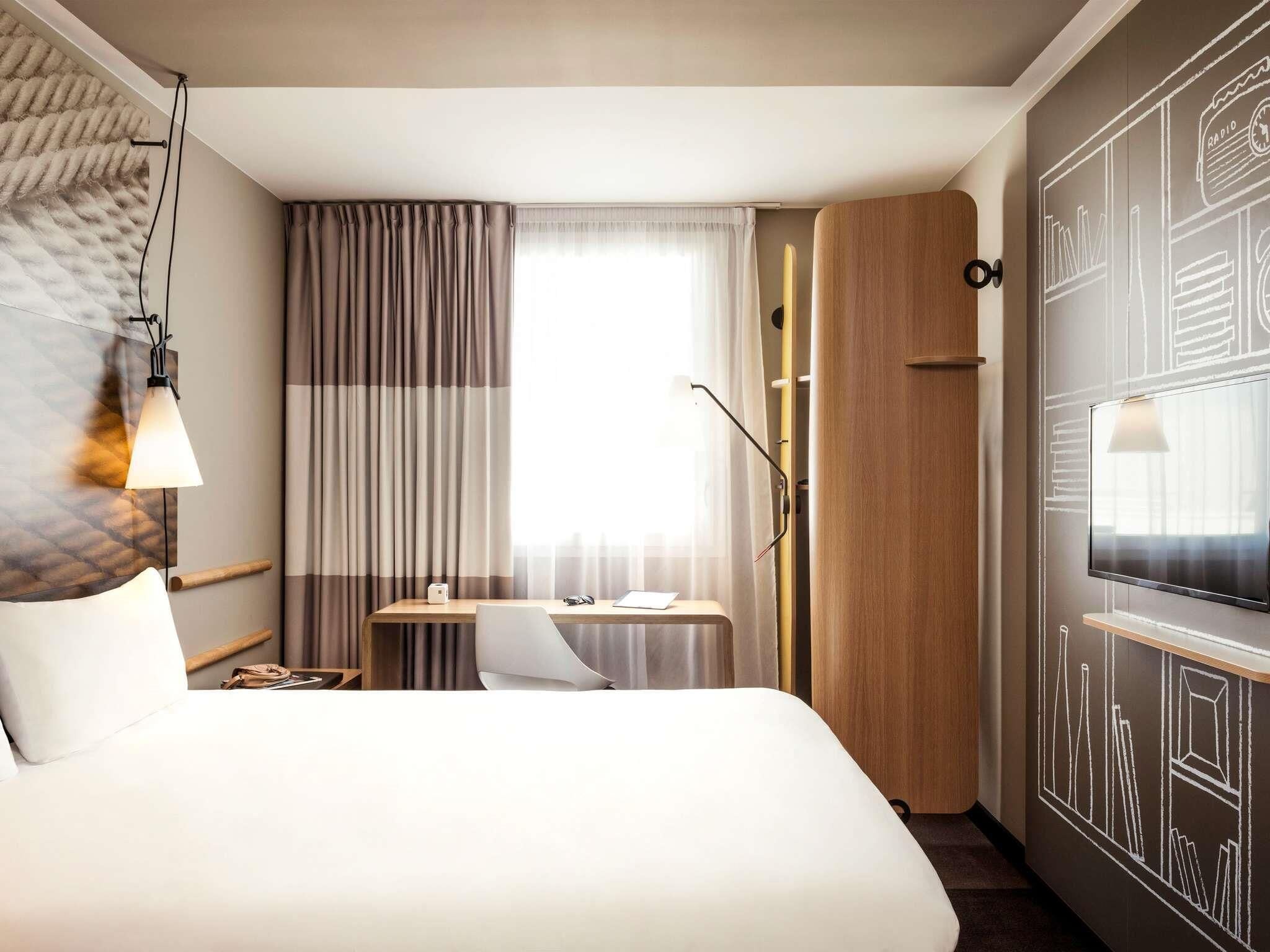 A hotel room with a bed, a desk, a chair, a television, and a large window with curtains.