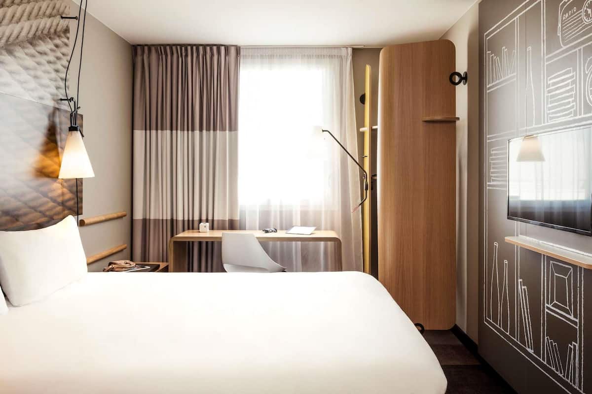 A hotel room with a bed, a desk, a chair, a television, and a large window with curtains.