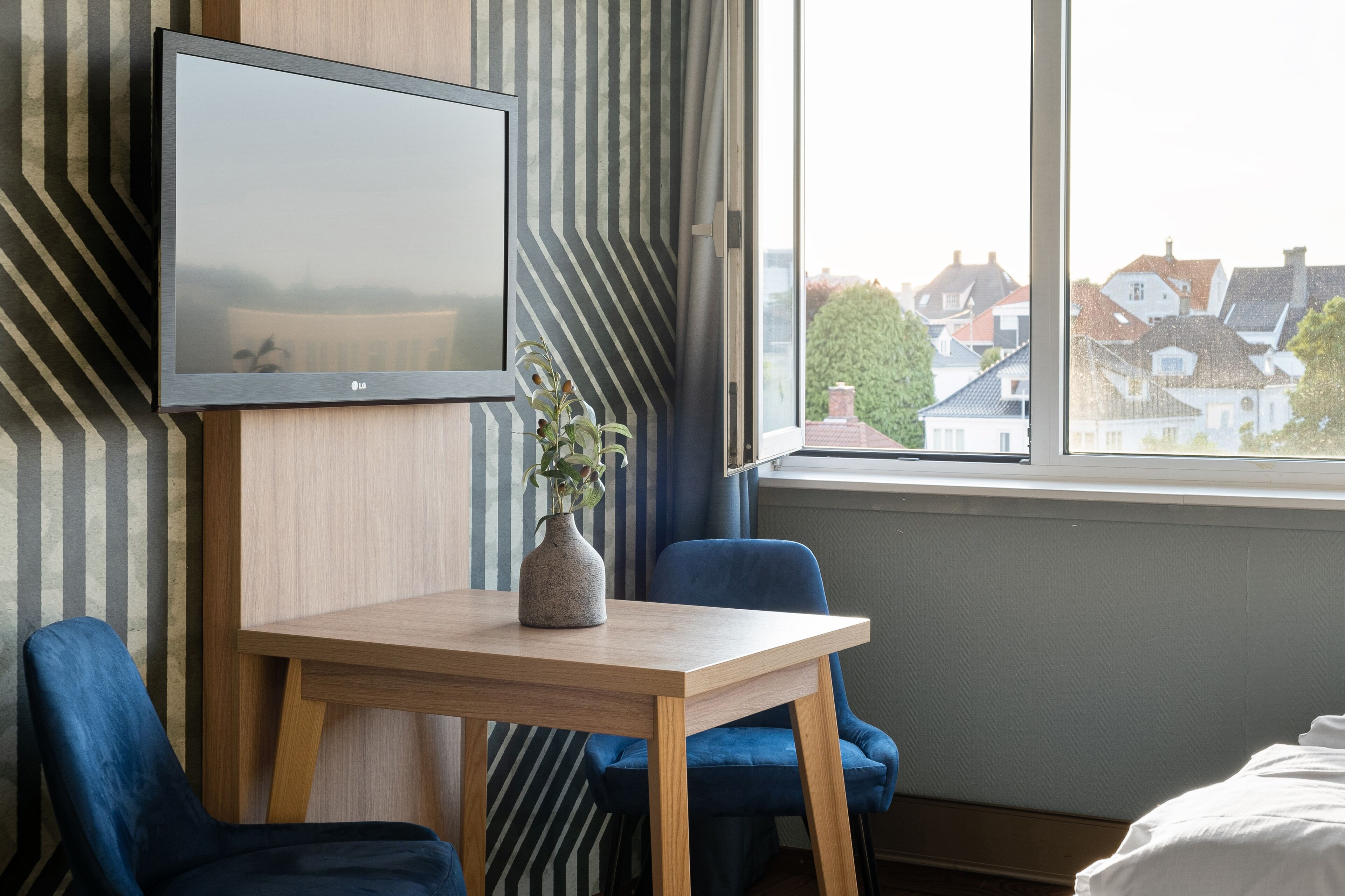A hotel room with a TV mounted on the wall, a wooden table with a vase, two blue chairs, and a large window offering a view of houses.