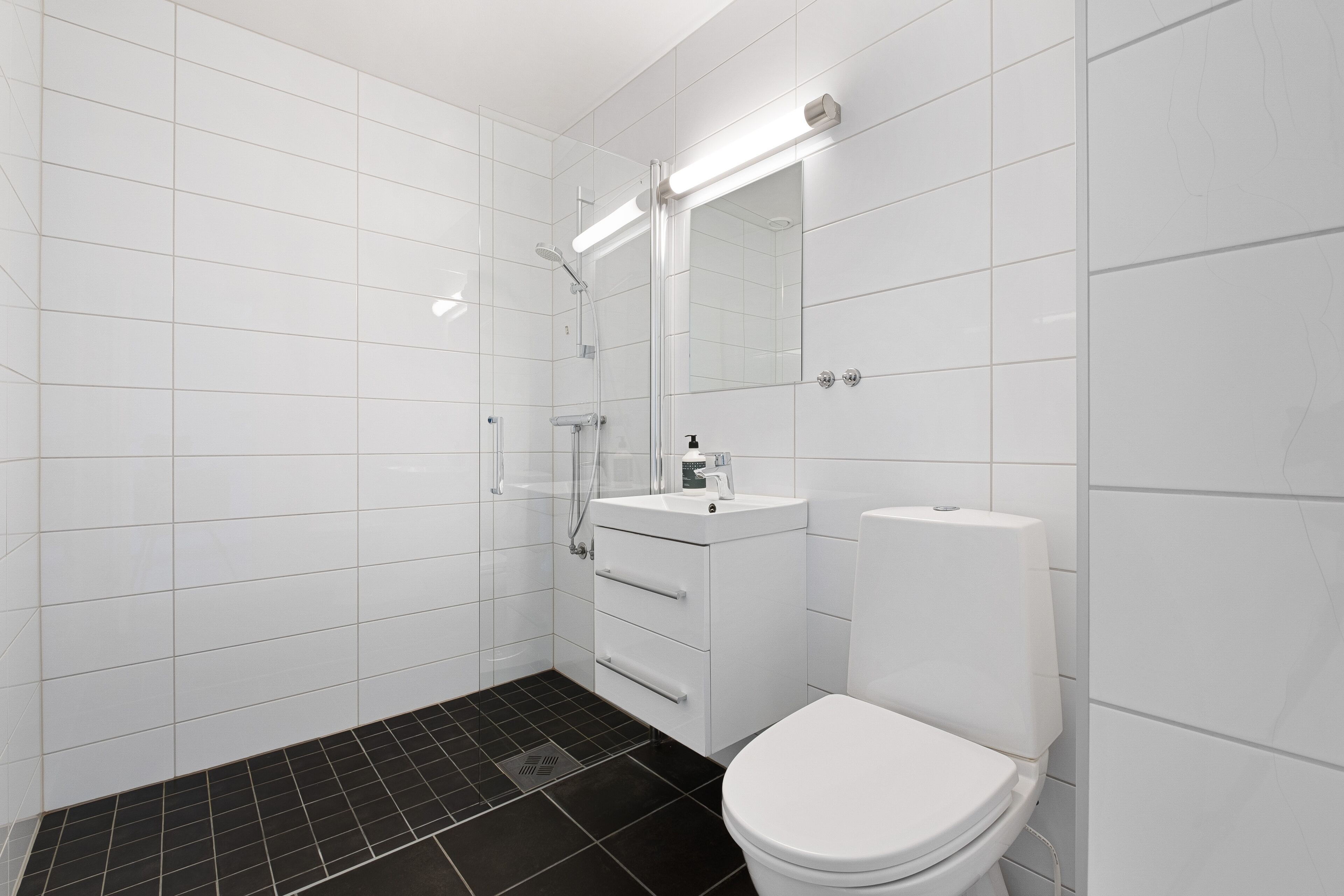 Un baño moderno con azulejos blancos, piso de ducha con mosaicos negros, inodoro blanco y un lavabo con espejo superior.