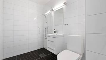 Un baño moderno con azulejos blancos, piso de ducha con mosaicos negros, inodoro blanco y un lavabo con espejo superior.