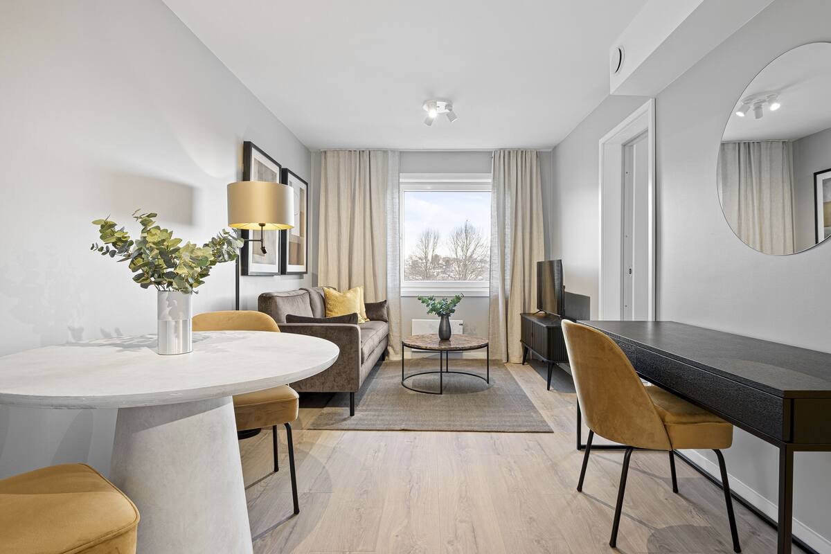 A modern hotel room with a round dining table, a sofa, a desk, and a large window with curtains.