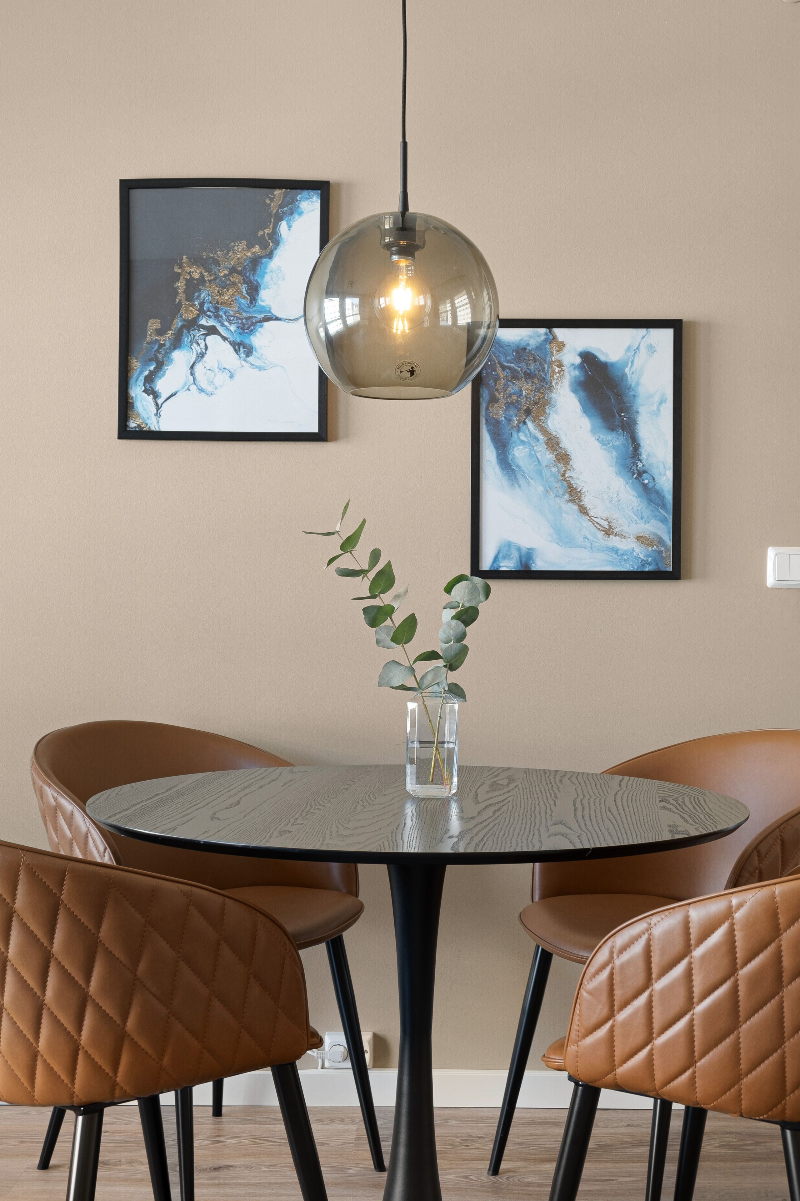 A dining area with a round table, four brown chairs, a glass vase with green leaves, and two abstract paintings on the wall.