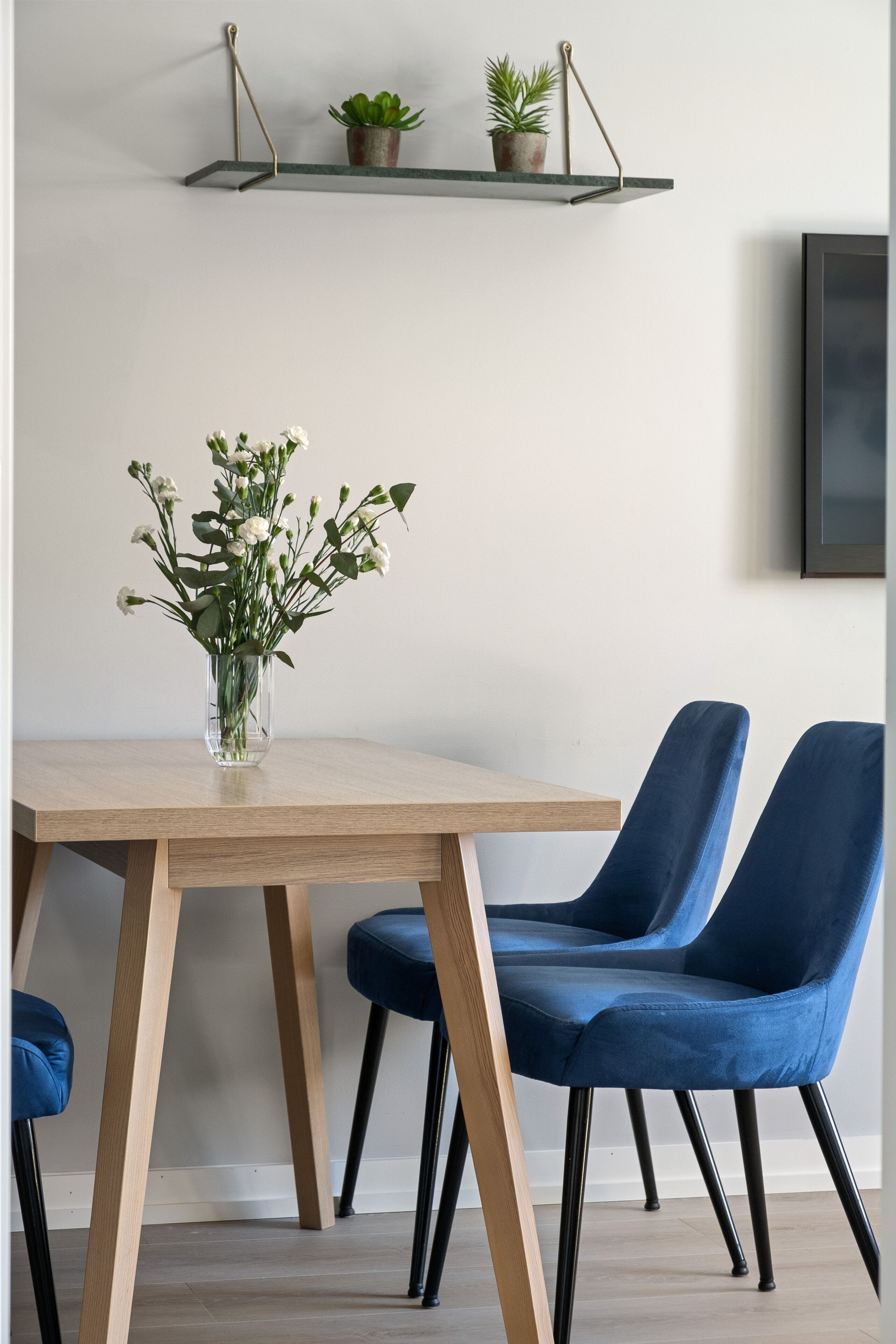 A dining area with a wooden table, two blue chairs, a vase with white flowers, and a shelf with plants.
