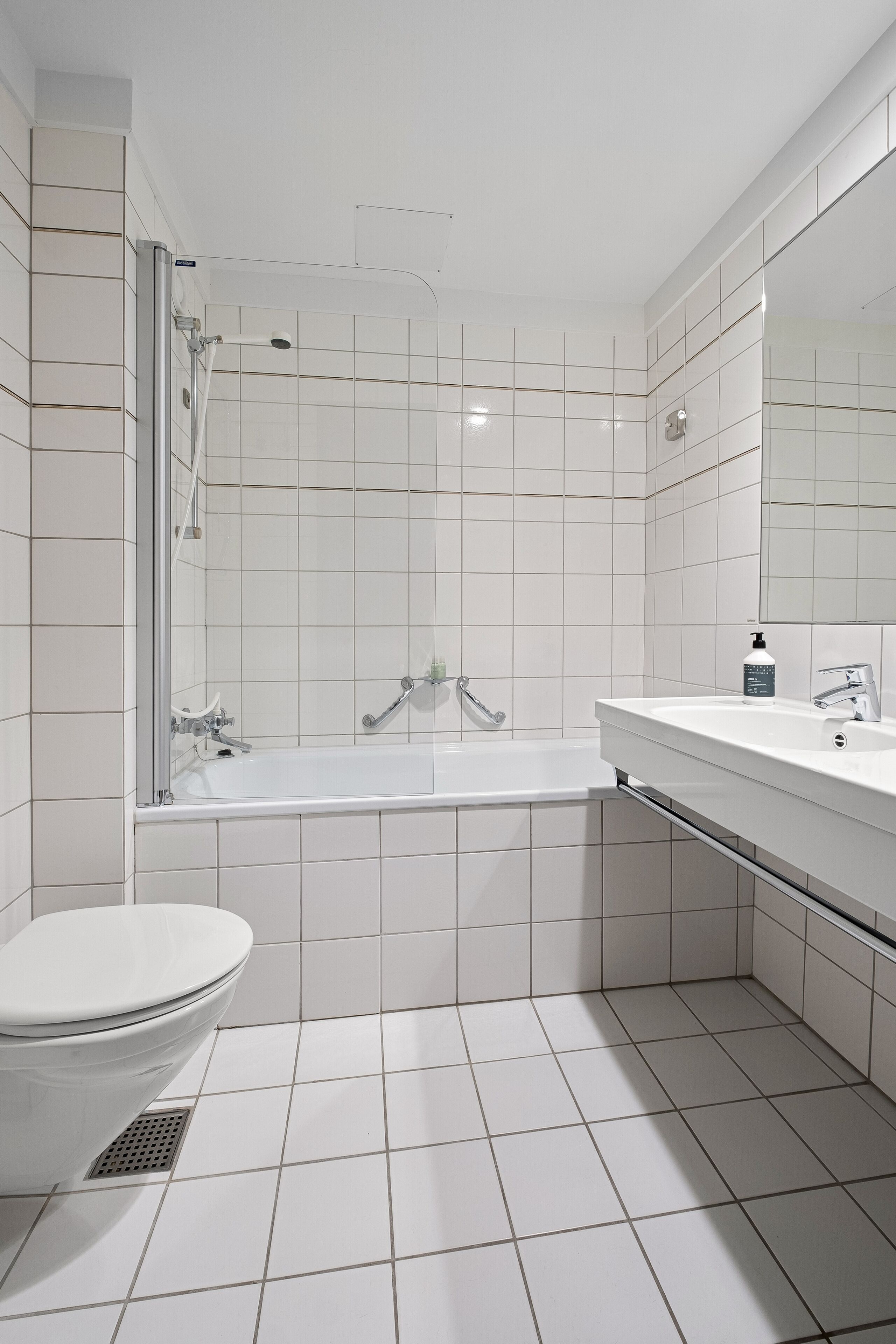 A modern bathroom with a white tiled floor, a white bathtub, a white toilet, and a white sink with a mirror above it.