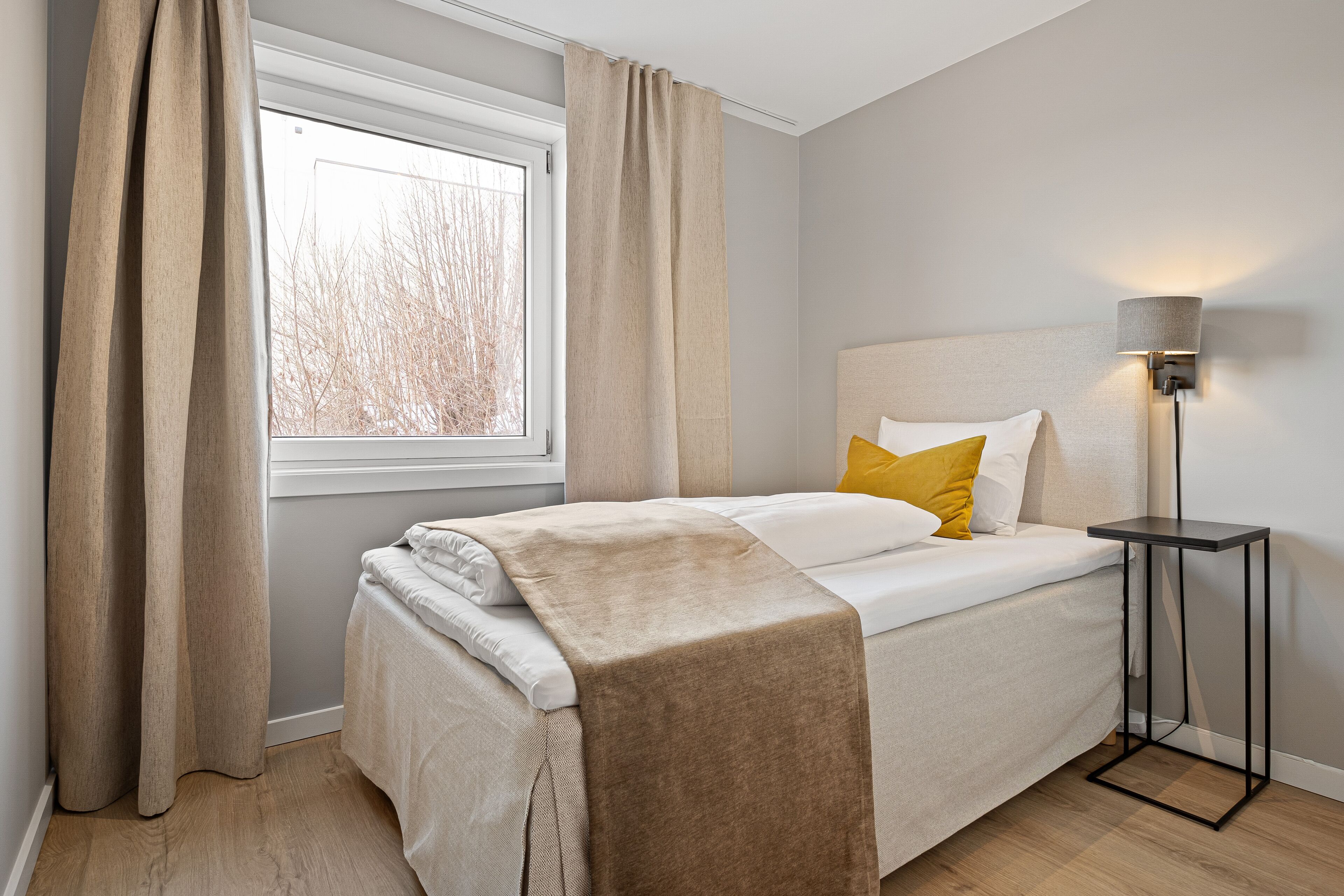 A neatly made bed with a beige blanket, a bedside lamp, and a window with curtains.