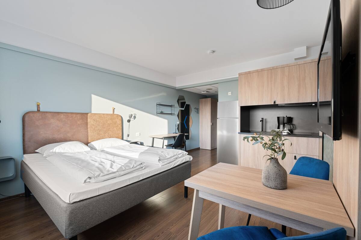 A modern hotel room with a bed, a desk, a chair, a small table with a vase, and a kitchenette area.