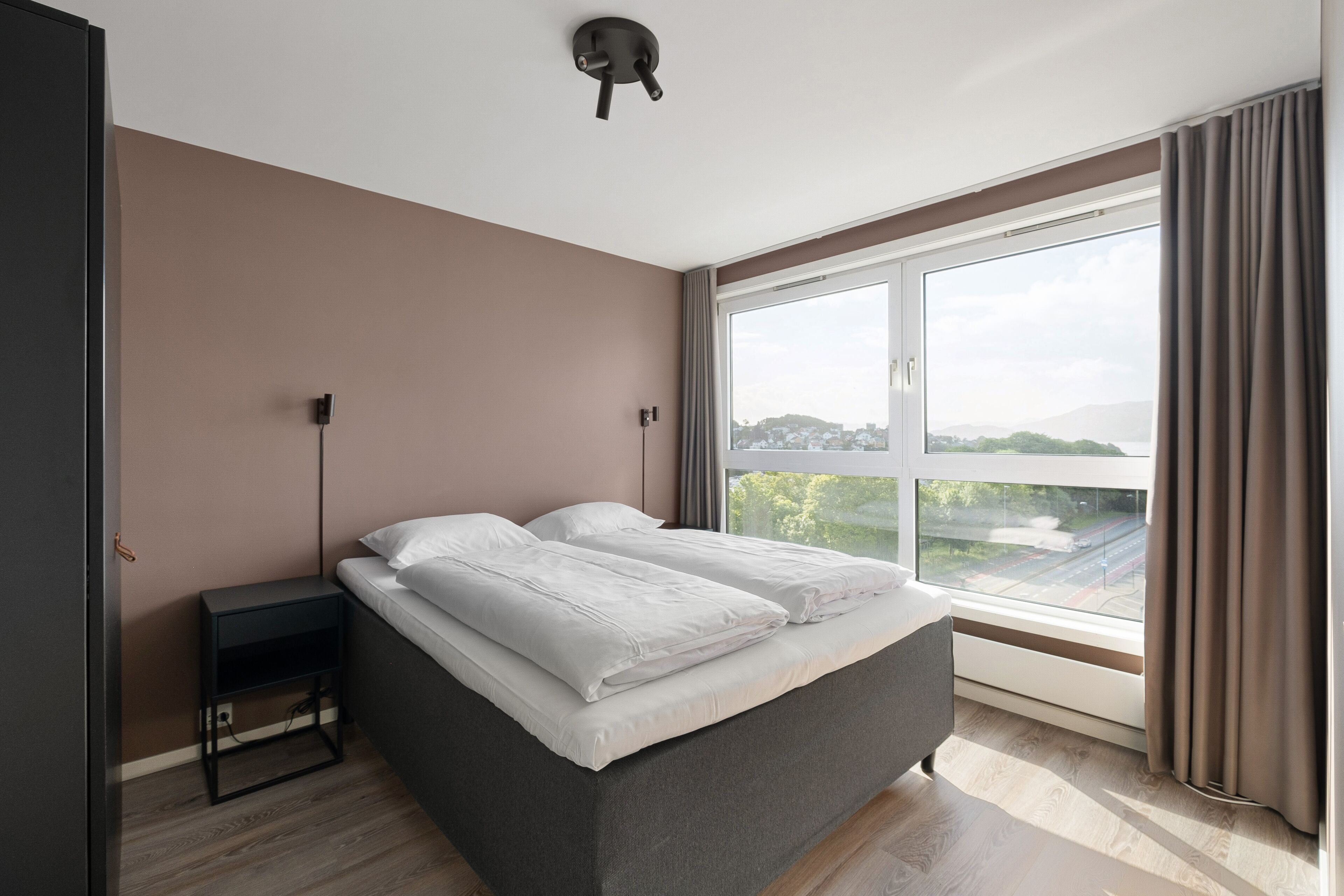 A modern bedroom with a large window, a bed, a bedside table, and a wall-mounted light fixture.
