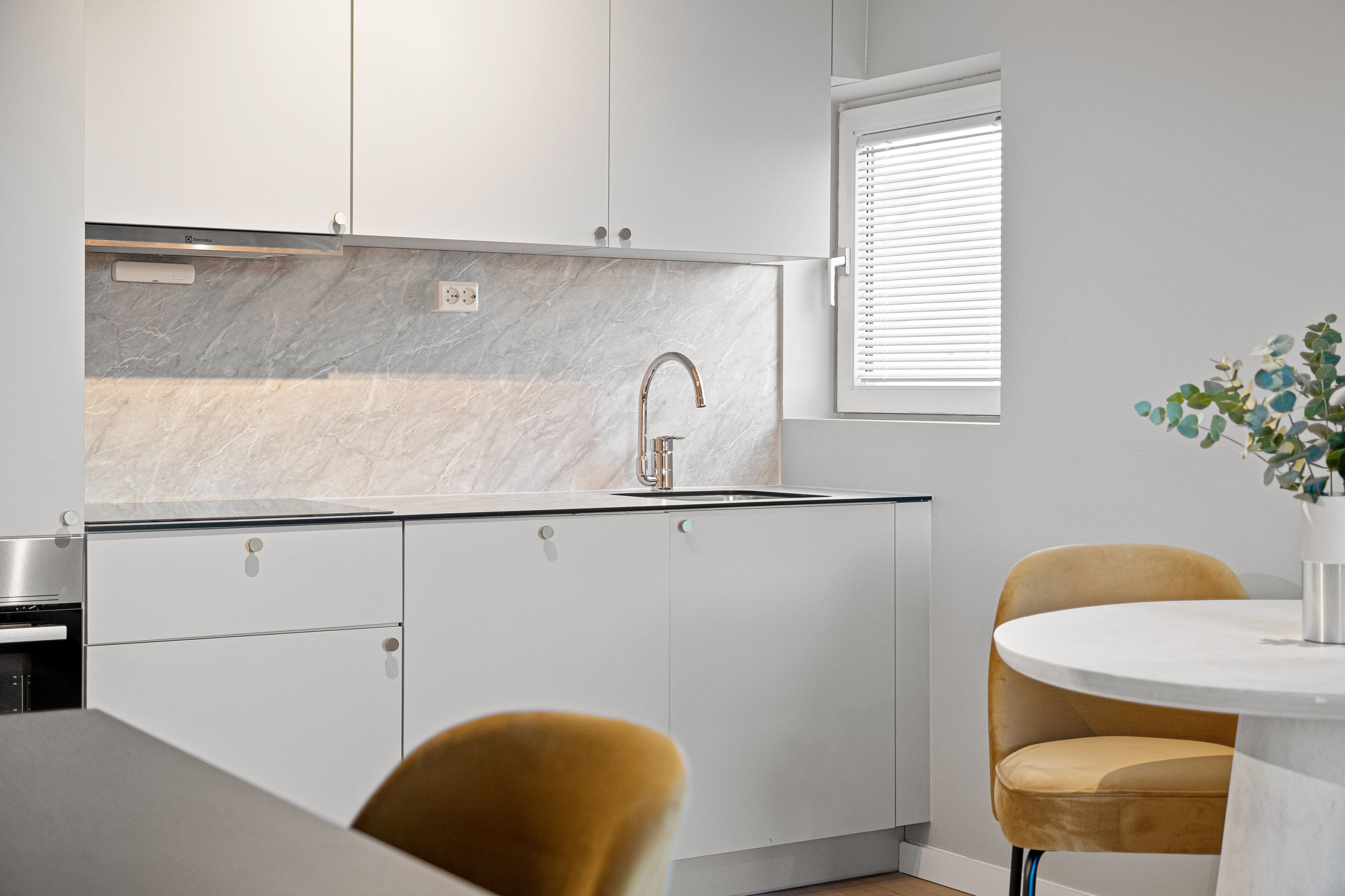 A modern kitchen with white cabinets, a marble backsplash, a stainless steel sink, and a round table with a yellow chair.