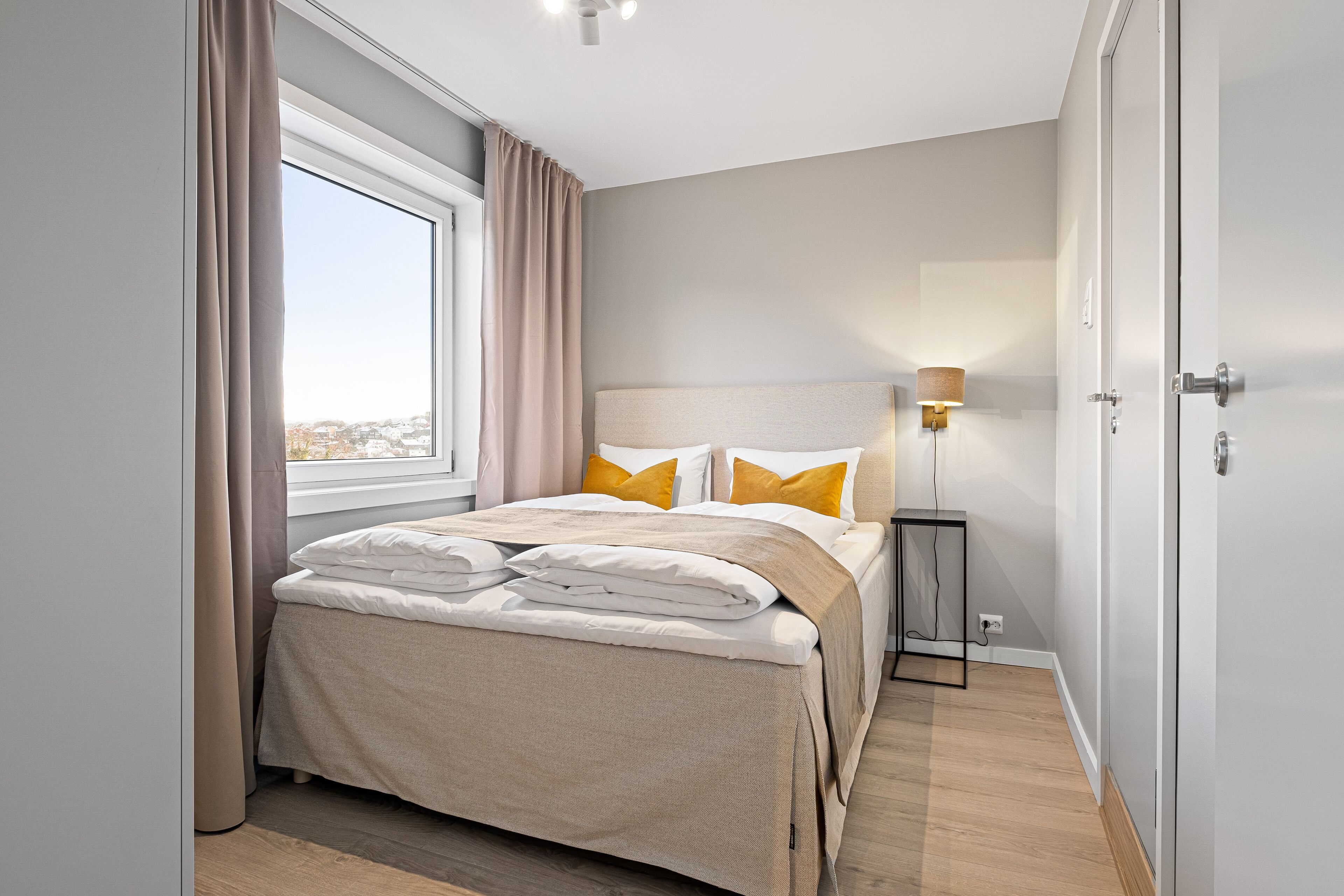 A modern bedroom with a large bed, a bedside table, a lamp, and a view of the city through a window.