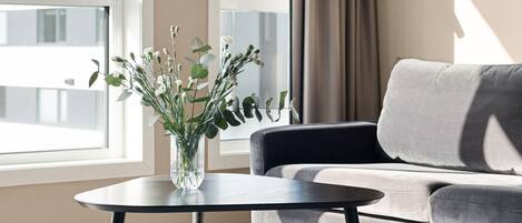 A modern living room with a sofa, a round coffee table with a vase of flowers, and large windows with curtains.