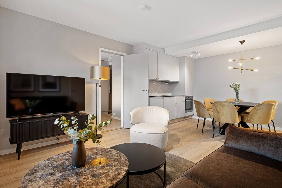 A modern living room with a flat-screen TV, a round coffee table, a sofa, and a dining area with yellow chairs.