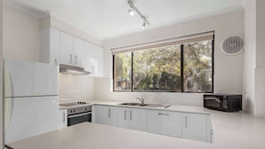 A modern kitchen with white cabinetry, a stainless steel refrigerator, a built-in oven, a microwave, and a large window providing a view of trees. - Best Western Geelong Motor Inn & Serviced Apartments (Geelong)