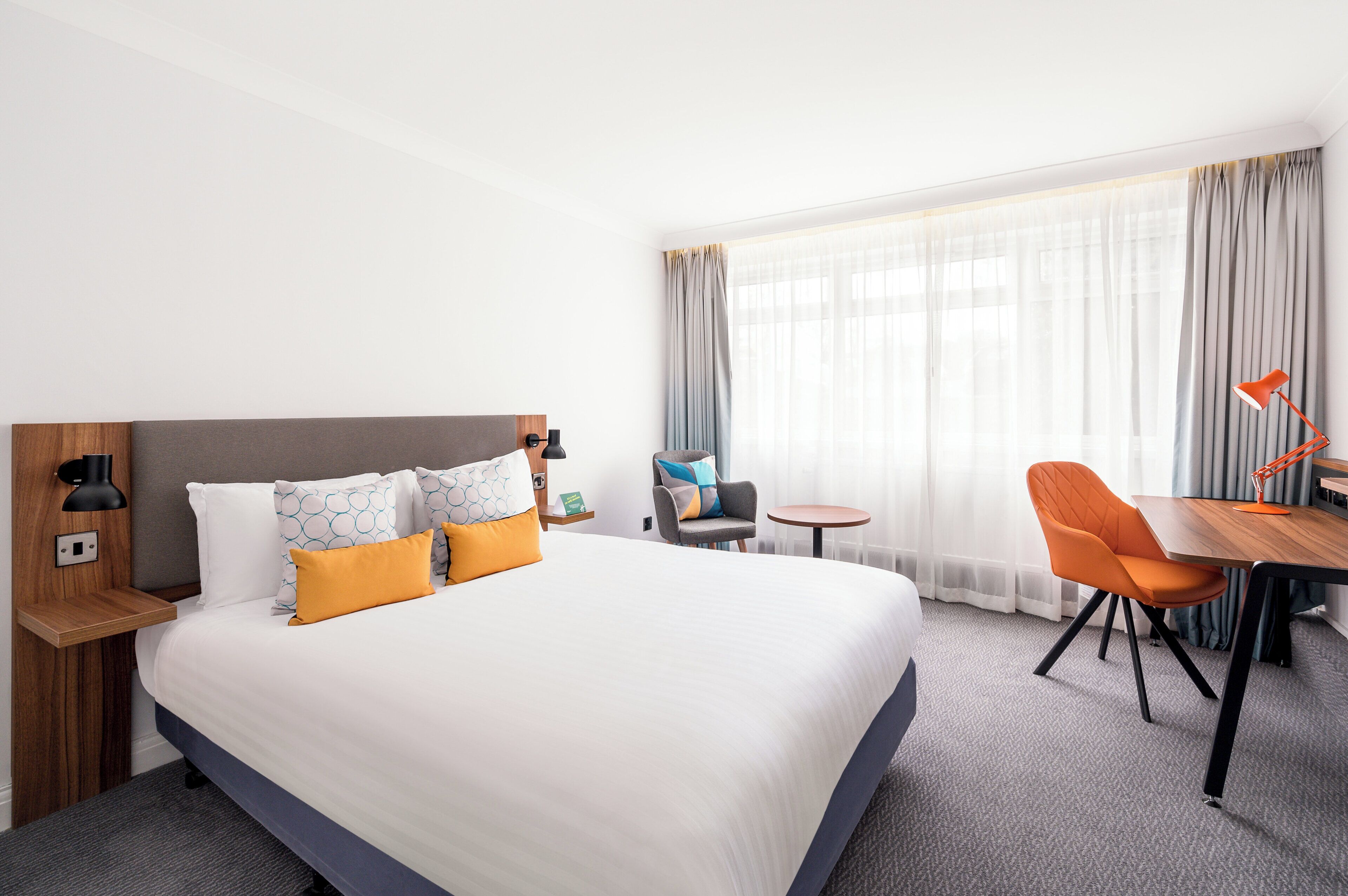 A hotel room with a large bed, a desk with a chair, a small table, and a lamp.