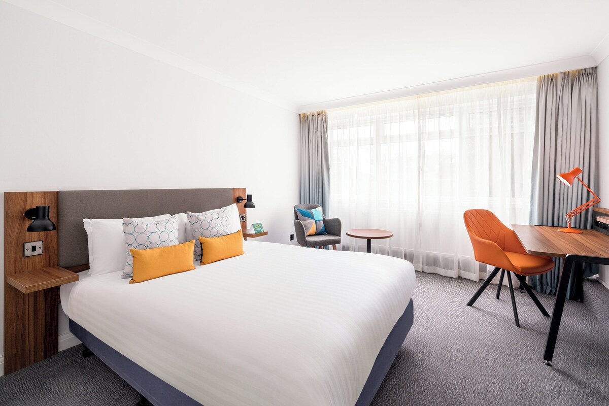 Habitación de hotel con una cama grande, un escritorio con una silla, una mesita y una lámpara.
