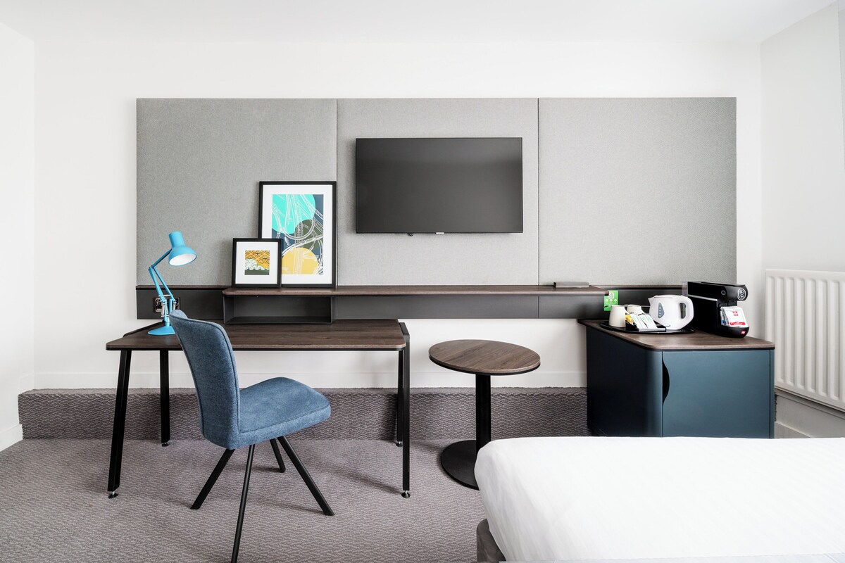 Una habitación de hotel moderna con una cama, un escritorio con silla, una mesita redonda pequeña, una cafetera y un televisor de pantalla plana montado en la pared.
