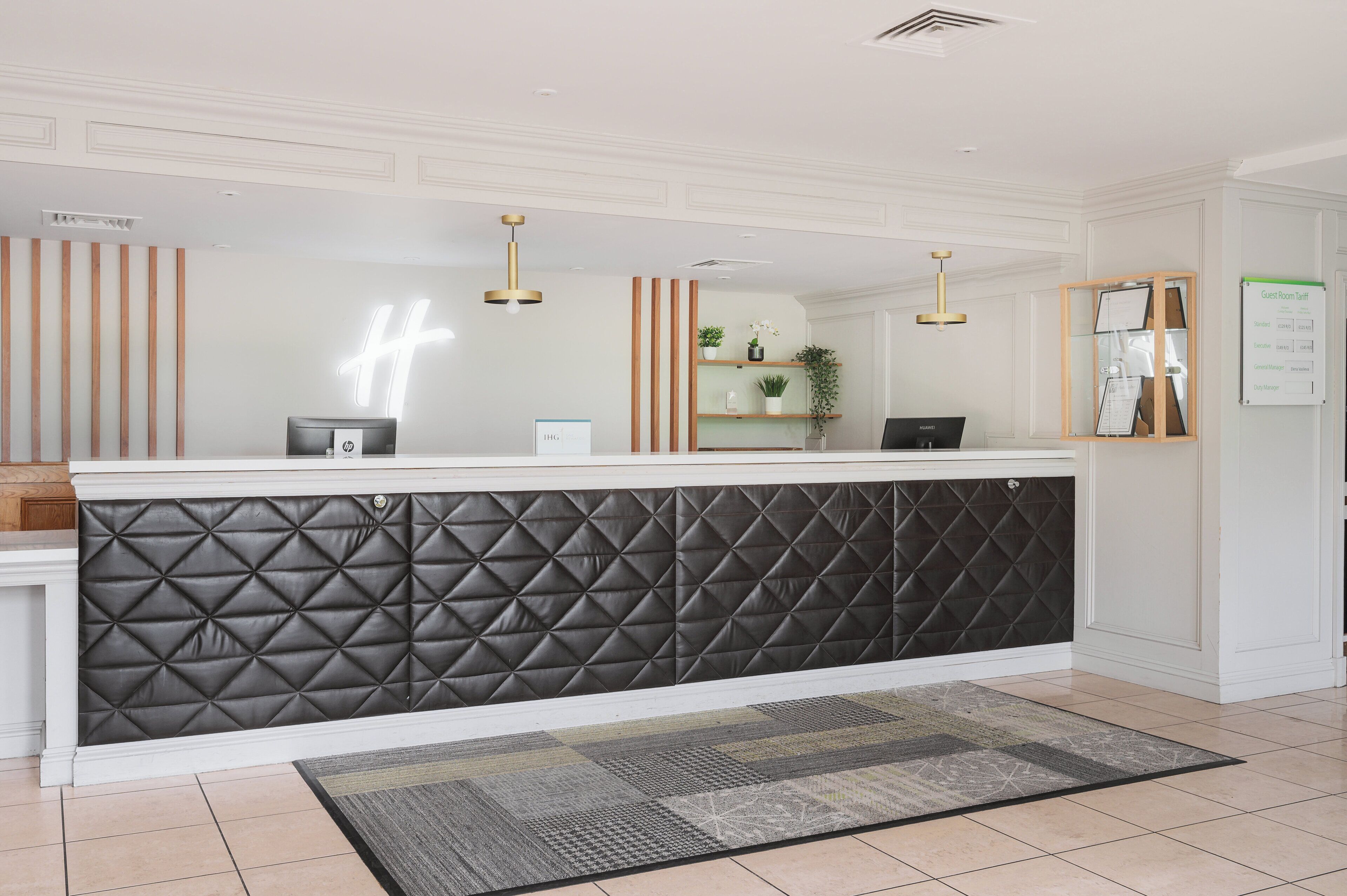 Ein Empfangsbereich mit schwarzer gepolsterter Wand, einer Rezeption, einem Computer, einer Pflanze und einer Tafel mit der Liste der Gästezimmer.