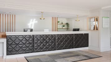 Ein Empfangsbereich mit schwarzer gepolsterter Wand, einer Rezeption, einem Computer, einer Pflanze und einer Tafel mit der Liste der Gästezimmer.