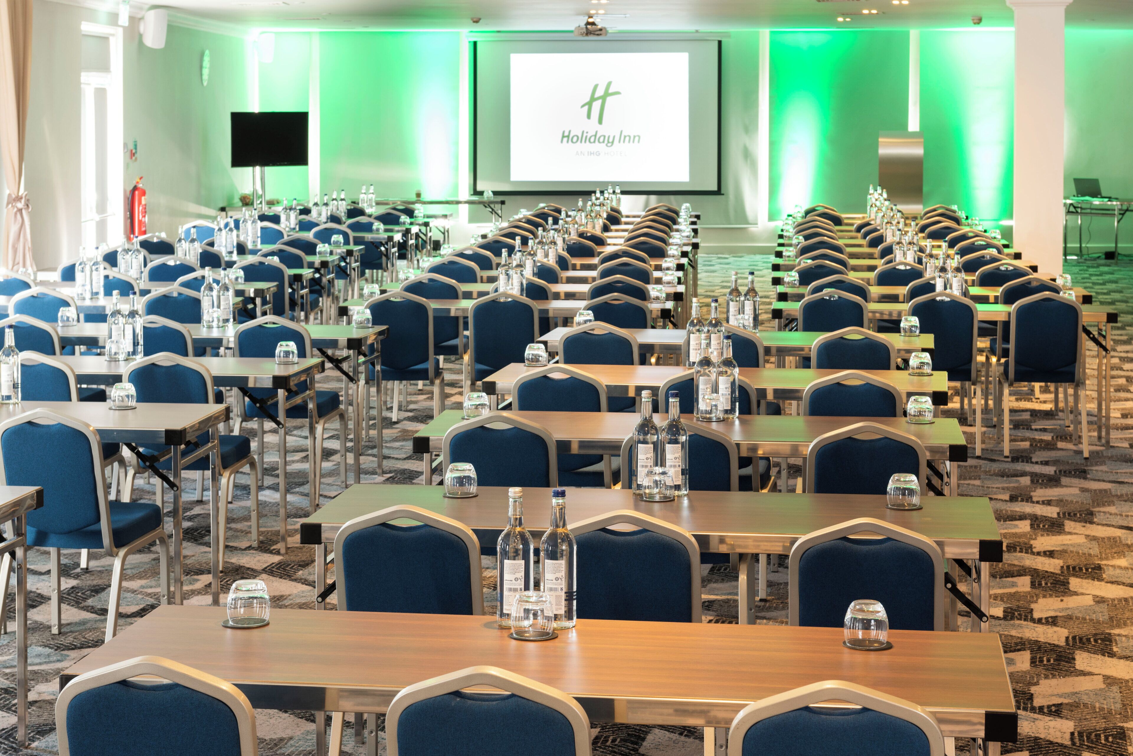 A conference room with rows of tables and chairs, a projector screen displaying 'Holiday Inn,' and a television on the wall.